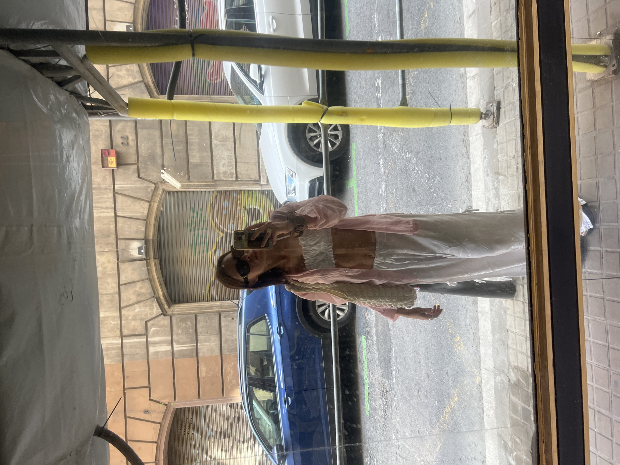 A young woman stands in front of a mirror, taking a selfie. She is wearing a white bandeau top, a pink open shirt, and white wide-leg pants. She has dark hair and is wearing sunglasses. A white textured bag hangs from her left shoulder. She is holding a smartphone in her right hand. The background shows a street scene in Barcelona, Spain. A blue SUV is parked on the left, and a white Audi SUV is parked on the right. The buildings in the background have traditional architecture with arched doorways and closed metal shutters, some of which are covered in graffiti. Yellow protective foam covers scaffolding poles in the foreground. The reflection shows the woman standing on a sidewalk with patterned tiles, and a street with grey asphalt and yellow lines. The scene appears to be captured during the daytime.