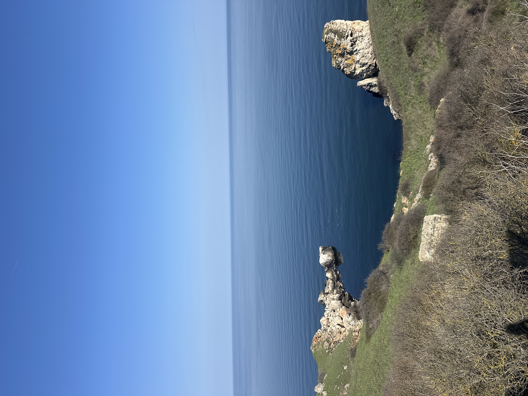 The image shows a vertical landscape of Kamen Bryag, Bulgaria. The top half of the scene is a clear blue sky transitioning into a lighter hue closer to the horizon where it meets the sea. The sea occupies the middle band of the image, appearing calm and deep blue.
The lower right portion is dominated by a rocky coastline covered with grass and sparse vegetation, including bare bushes and a patch of green grass. A series of rocky outcrops are seen jutting out into the sea. One prominent rock formation is composed of stacked rocks. A flat, square concrete or stone slab is seen amidst the vegetation.