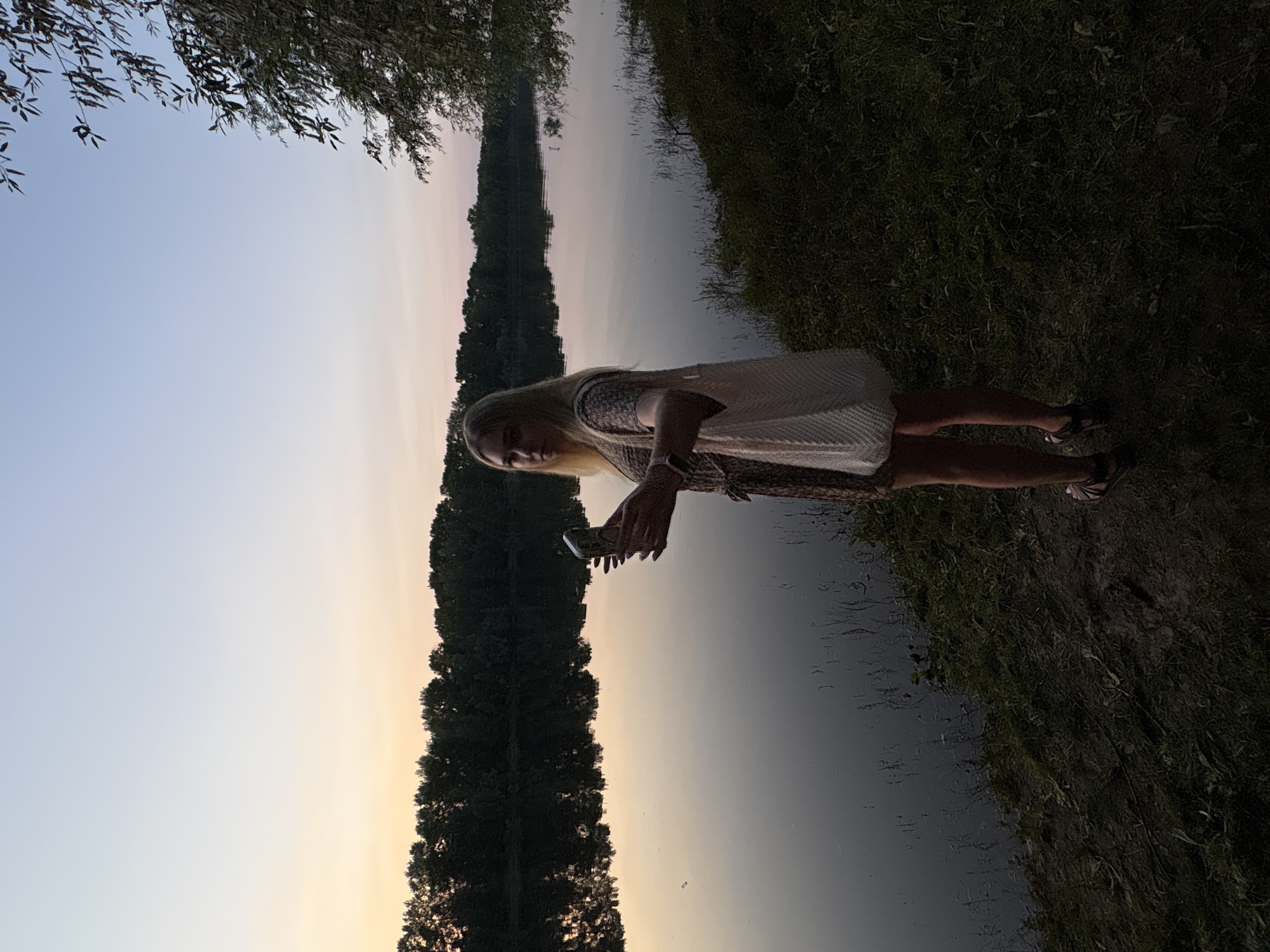 A young blonde woman stands on the grassy bank of a river at dusk, taking a selfie with her phone. She is wearing a patterned dress and sandals, and carrying a white mesh bag over her shoulder. The river is calm and reflects the pale pink and blue hues of the sky. A line of trees can be seen across the water. The scene appears to be a natural setting, possibly a park or embankment, in the city of Chaikovsky, Russia.