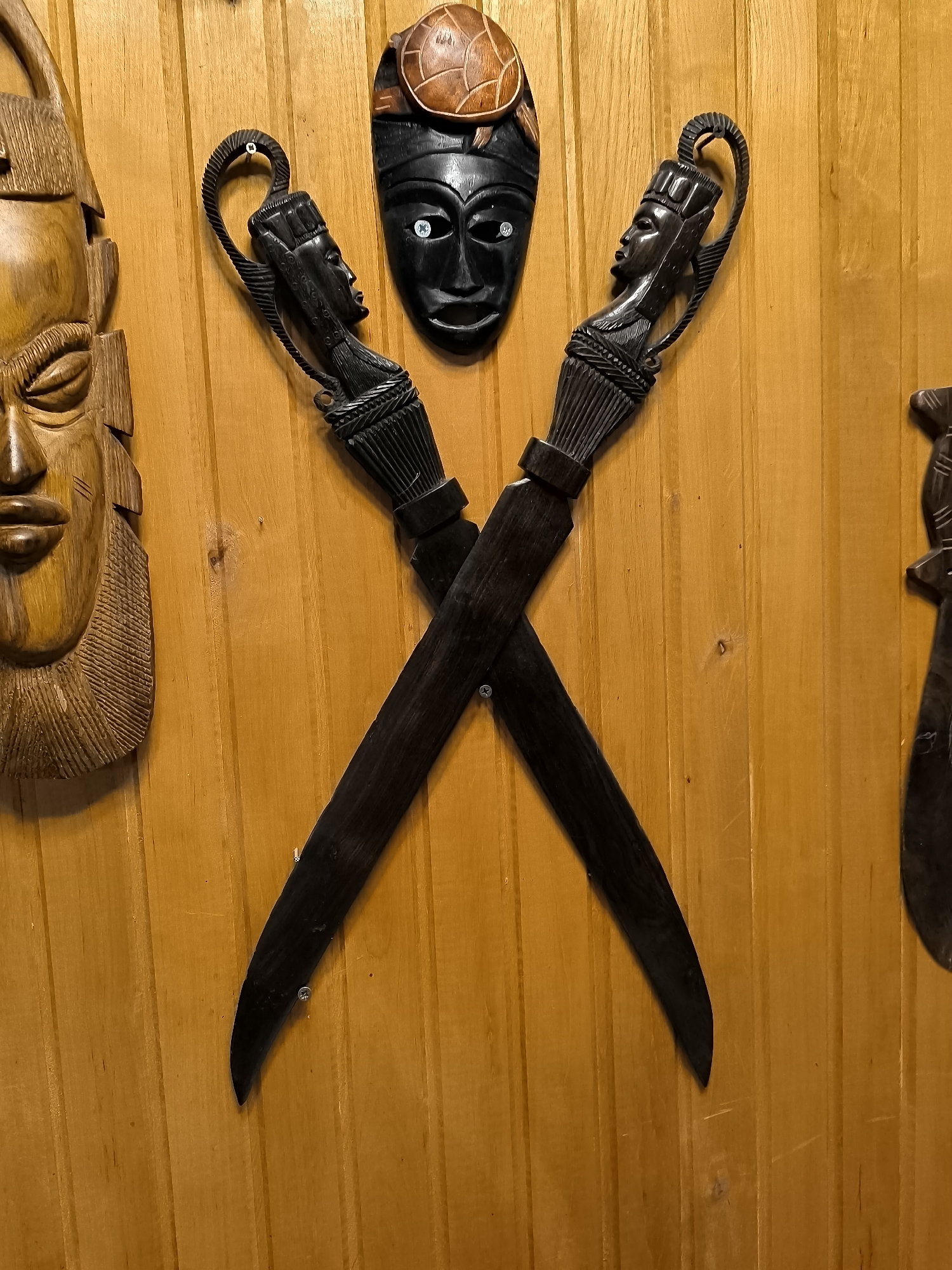The image shows a collection of African tribal art objects mounted on a wooden wall. In the center, two dark, carved wooden daggers with sculpted handles are crossed. Each handle depicts a stylized human head. Above the crossed daggers, a black carved mask with inlaid white and blue circular eyes is displayed, topped with a carved brown turtle shell. To the left of the central display, a large, light brown carved wooden mask with detailed facial features, including textured hair and segmented teeth, is visible. To the far right, a sliver of another dark carved object, possibly a mask or weapon, can be seen. The wall is constructed of vertical wooden planks with a warm, orange-brown hue. The objects are secured to the wall with visible screws. The scene is indoors, with the lighting casting soft shadows that highlight the textures of the carvings. The image does not provide any information about the city of Žiaukos, Lithuania.