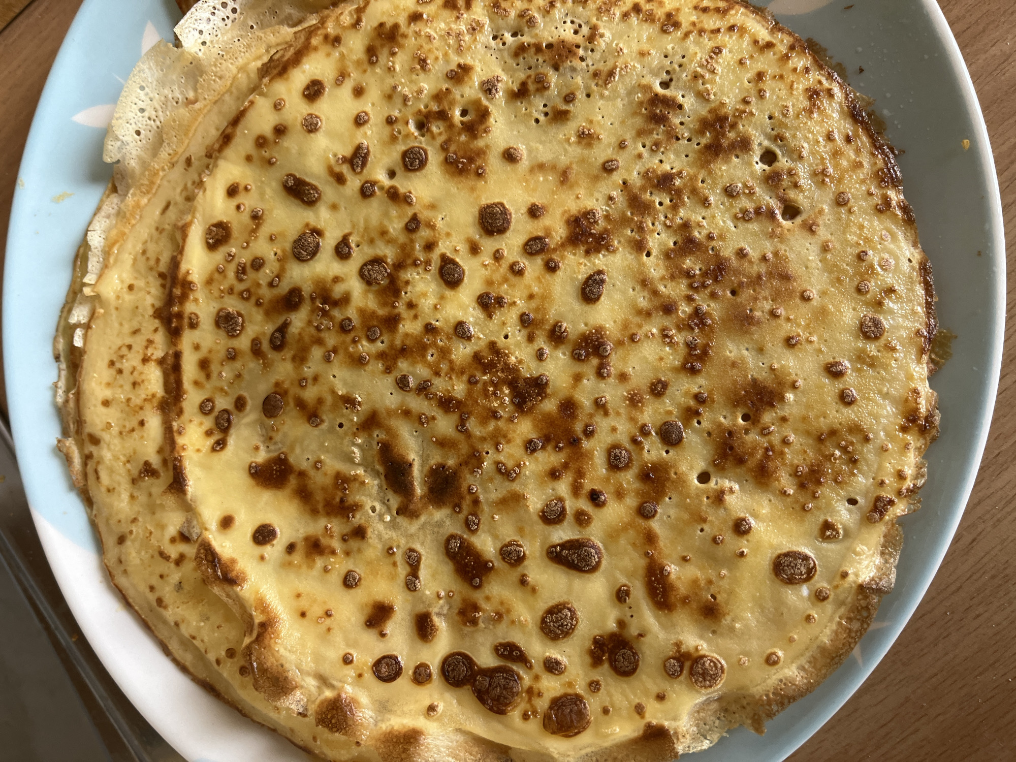 A stack of thin pancakes or crêpes sits on a blue-rimmed plate. The pancakes are golden brown with darker brown spots and edges. The top pancake is the most visible, covering the other pancakes in the stack. The plate rests on a wooden surface. The image does not contain evidence to confirm location in Boreti, Montenegro.