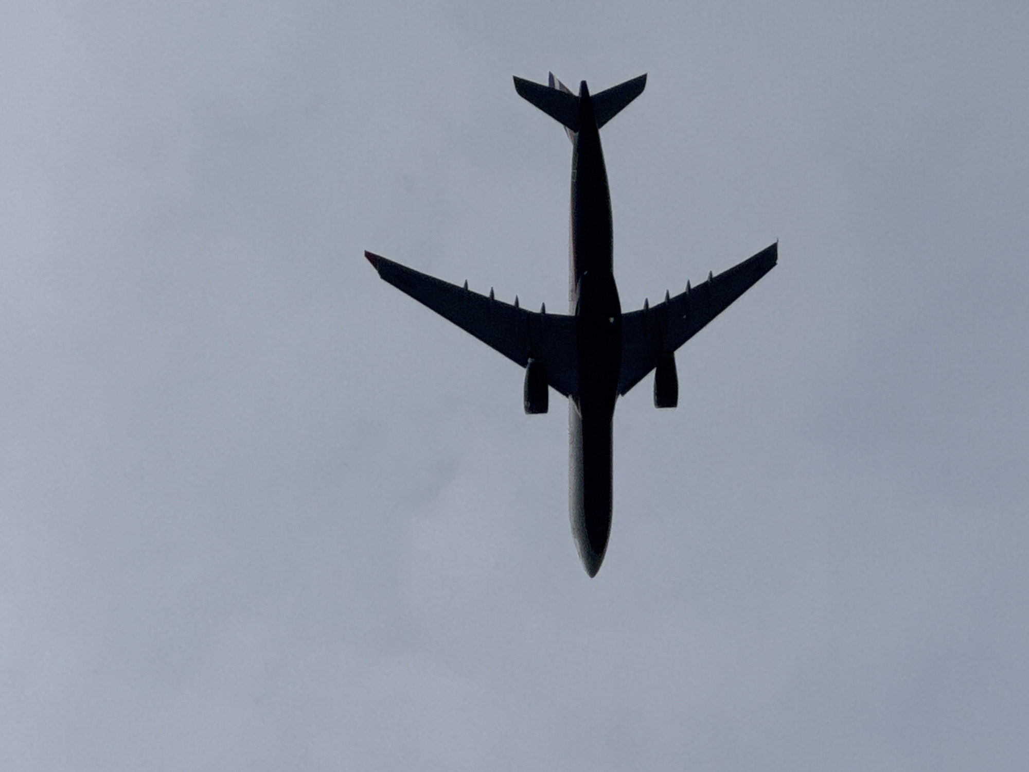 A dark silhouette of an airplane is captured from below, flying against a hazy, overcast sky. The aircraft is viewed from a low angle, with its wings spread wide and its engines visible under the wings. The tail of the plane is visible, pointing upwards. The sky is a uniform pale gray, with no discernible features. The image does not contain any information to suggest the location in the city of Kolpino, Russia.