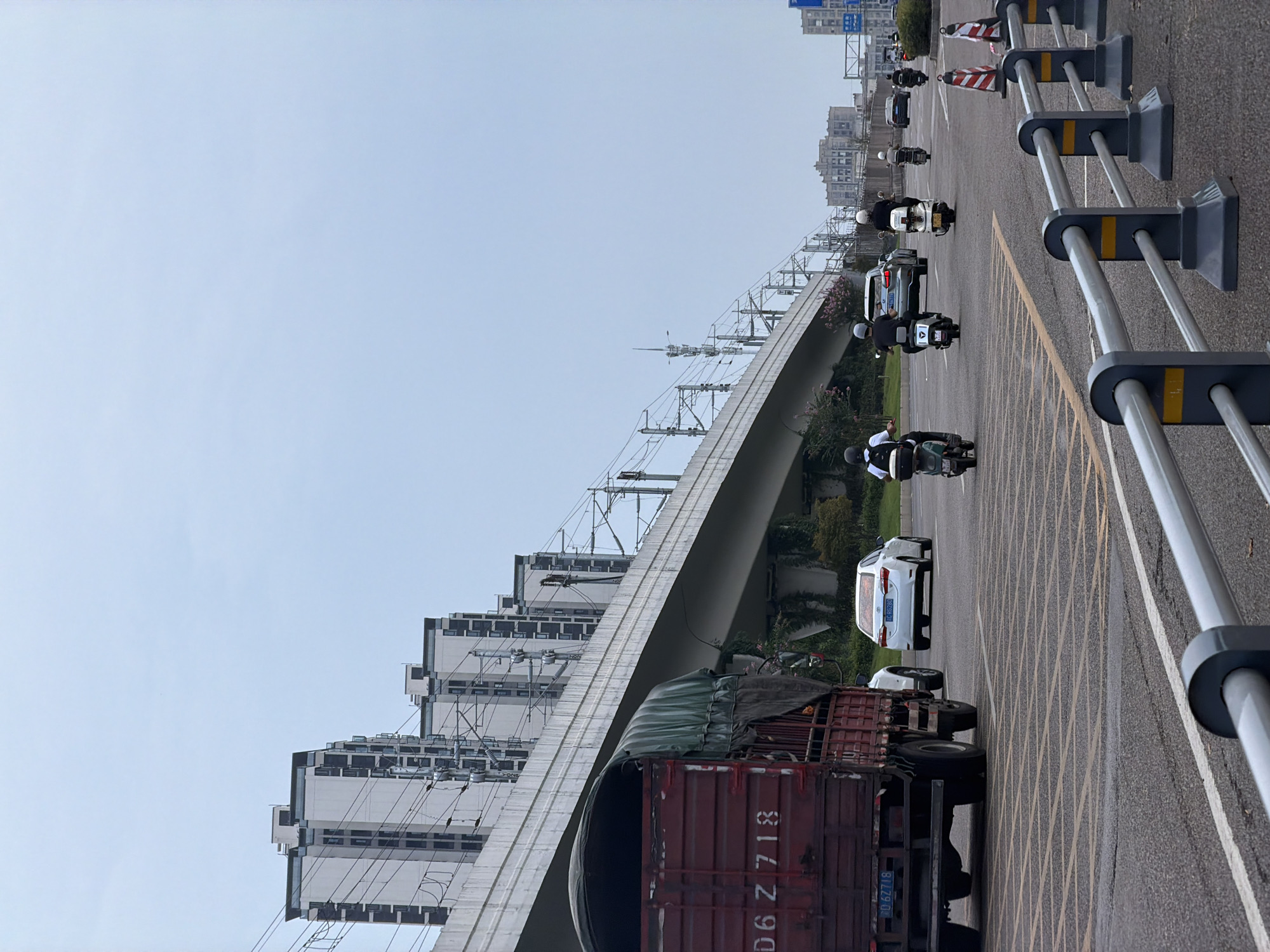 The image captures a street scene in the city of Jiangjin, China, under an overcast sky. In the foreground, a metal railing with dark gray bases and yellow accents separates the viewer from the road. The road surface is asphalt, marked with yellow grid lines. A red truck with a green tarp is prominently positioned on the left, displaying white characters that read "D6 Z 718". Behind the truck, a white sedan with license plate "2 RG280" is visible.
Several individuals are riding on scooters in the middle ground, heading away from the viewer. One scooter rider in the foreground is wearing a white helmet and a black shirt, and appears to be looking back. Other scooter riders are further ahead, also dressed in casual attire. In the background, a raised concrete structure, likely an elevated road or railway line, runs horizontally across the frame, supported by concrete pillars. Buildings with multiple stories, characterized by white facades and dark windows, are lined up along the elevated structure. Utility poles with complex wiring are also visible alongside the elevated structure. Further in the distance, more buildings and streetlights are discernible. The overall atmosphere is one of urban transit and everyday life.