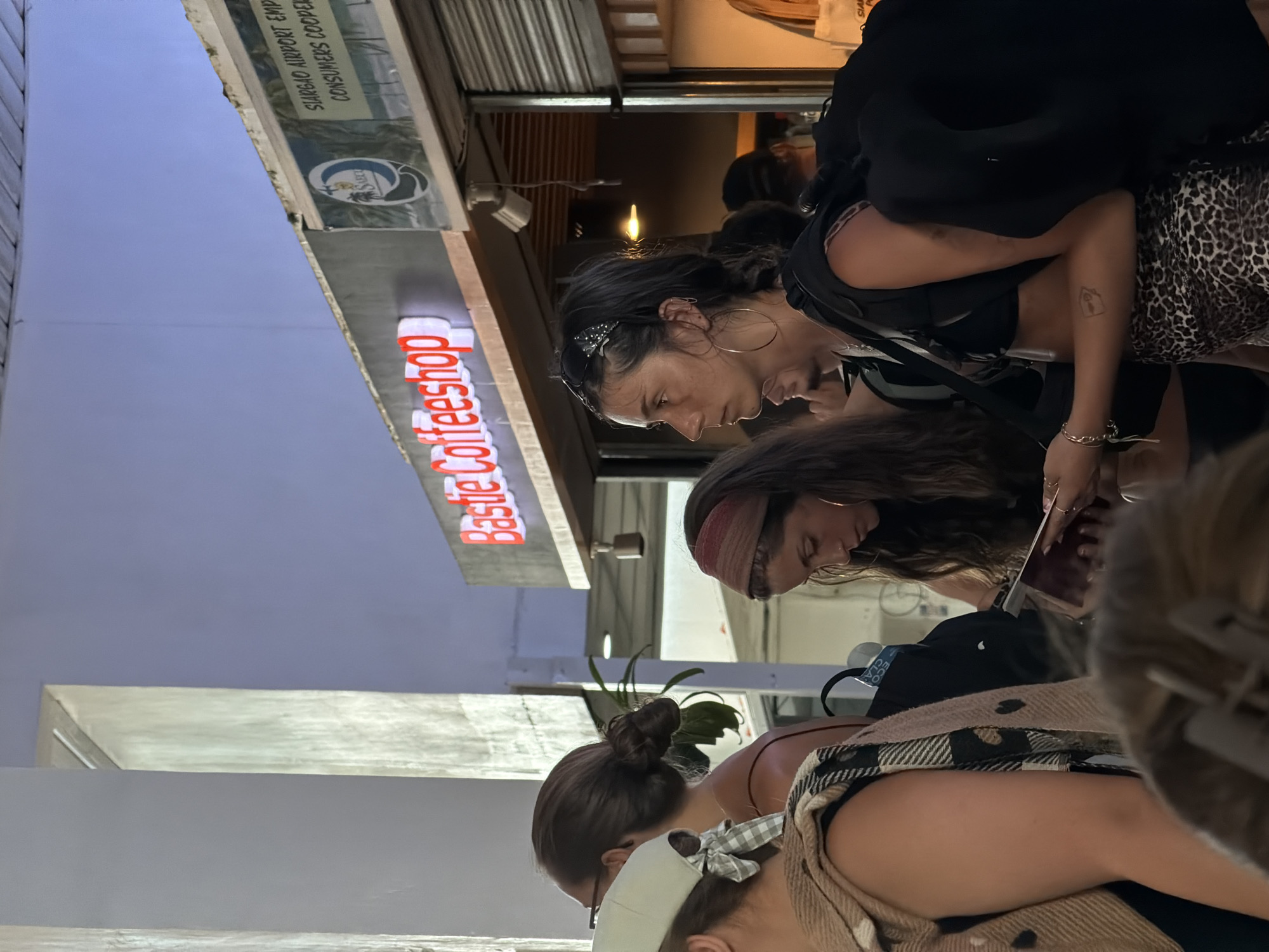 The image shows three women standing on a street outside "Bastie Coffeeshop" in Del Carmen, Philippines. The cafe sign is red. One woman wears hoop earrings, a headband, and has a tattoo on her arm. Another wears a red headband, and the third is wearing a plaid scarf, a hat, and has their hair up in a bun. The lighting suggests it is daytime. A sign indicates this is the location of the Siargao Airport Employees Consumers Cooperative.