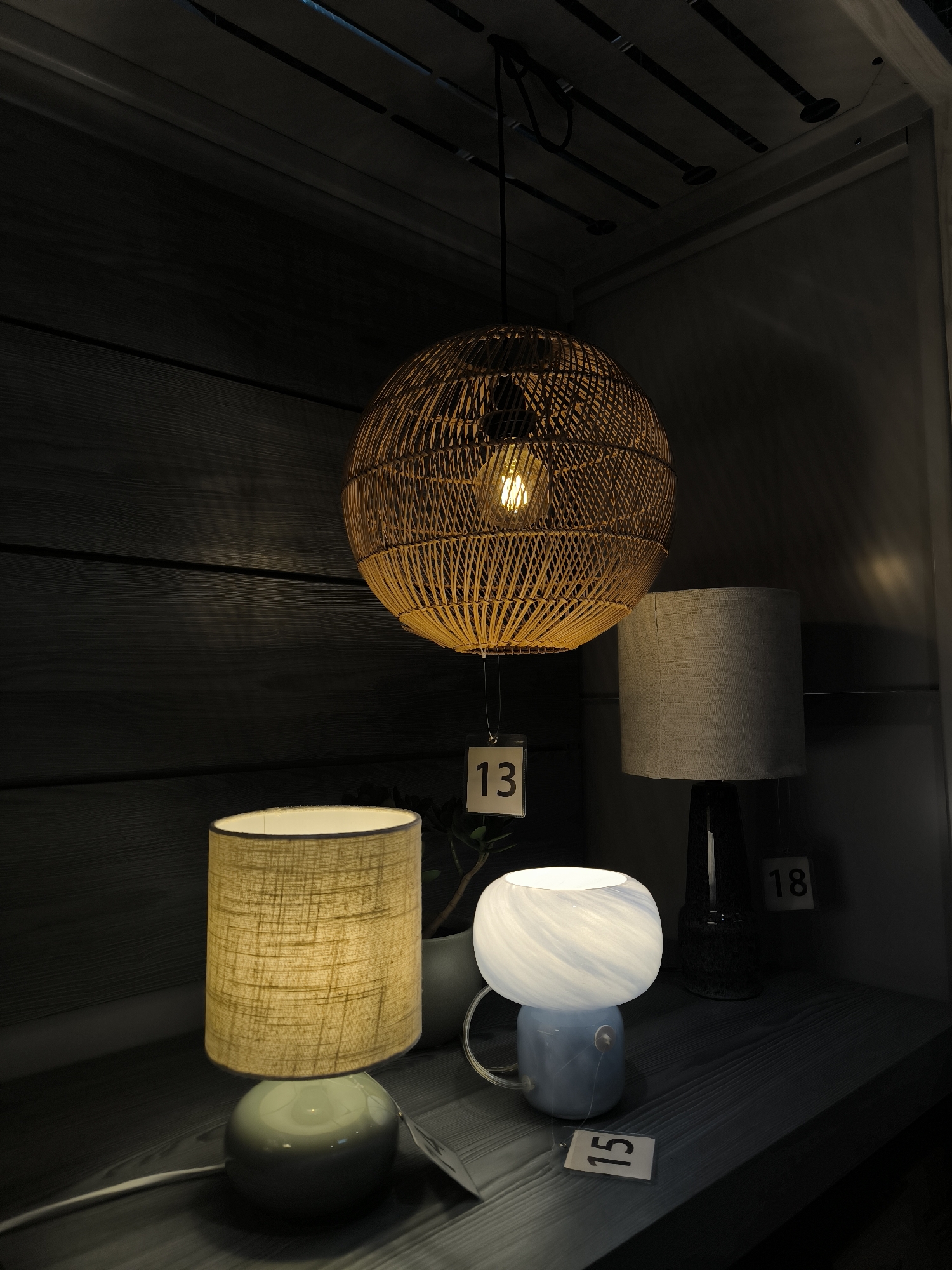 The image displays an indoor scene featuring a collection of illuminated lamps arranged on a dark wooden shelf or table, set against a dark wooden paneled wall. In the upper center, a large, spherical pendant lamp, constructed from woven wicker, hangs from the ceiling, emitting a warm, bright glow from its exposed bulb.
On the table below, three different table lamps are visible. To the left, a cylindrical table lamp with a textured, light beige fabric shade sits atop a round, glossy teal base. Next to it, in the center, is a smaller, distinctive lamp with a light blue base and a translucent white glass shade that features swirling patterns, also emitting light. Partially visible on the right, another table lamp with a grey fabric shade and a dark base is present. A small, dark-potted plant is subtly placed between the left and center table lamps.
The overall lighting in the scene is dim, with the lamps providing the primary illumination, creating a cozy and inviting atmosphere. The wooden surfaces have visible grain, adding texture to the setting. Above the pendant lamp, various dark pipes or fixtures are visible against a lighter ceiling.
Several white tags with numbers are visible. A tag reading "13" hangs below the wicker pendant lamp. Another tag with "15" is placed on the table in front of the blue and white table lamp. A third tag, showing "18", is partially visible behind the grey-shaded table lamp on the right. The scene appears to be a product display, likely in a store or showroom, showcasing different lighting options.