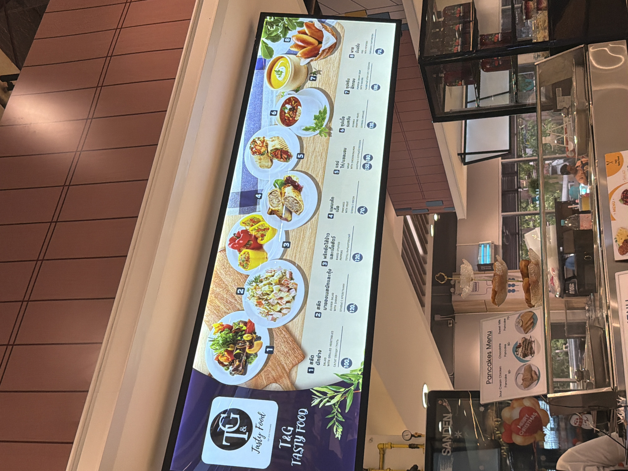The image shows a digital menu board displaying various food items at "T&G Tasty Food" in Ban Bang Rak, Thailand. The menu is presented in Thai and English, with numbered items featuring images of the dishes. Items include salads, baked stuffed peppers, wraps, and soups, with prices indicated. Below the digital menu, there is a physical "Pancakes Menu" board and display cases containing food. People are visible in the background, suggesting a restaurant or food court setting. The overall environment appears to be an indoor establishment with bright lighting.