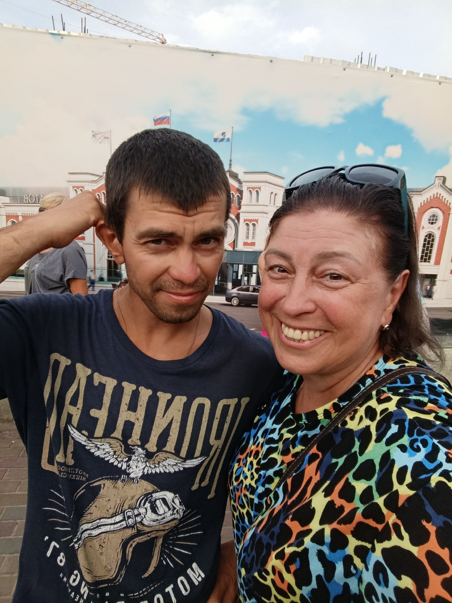 A medium shot captures two individuals posing for a selfie outdoors, with a backdrop resembling a painted mural of buildings and a cloudy sky. The person on the left is a man with short, dark hair and a beard, wearing a dark blue t-shirt with a graphic design. He is touching his ear with his right hand. The person on the right is a woman with dark hair pulled back and sunglasses perched on her head. She is wearing a brightly colored, patterned top with black, blue, green, yellow, and orange spots. She is smiling widely. In the background, there are elements of what appears to be a construction site with a crane, and the mural depicts classical-style buildings, possibly a train station or public building, with flags. A dark car is visible parked in front of one of the depicted buildings. The ground in the foreground appears to be paved with brick. The overall impression is a casual, daytime photograph in an urban setting. While the specific location in Saratov, Russia is not explicitly indicated by any visual cues, the presence of Russian flags on the mural suggests a Russian context.