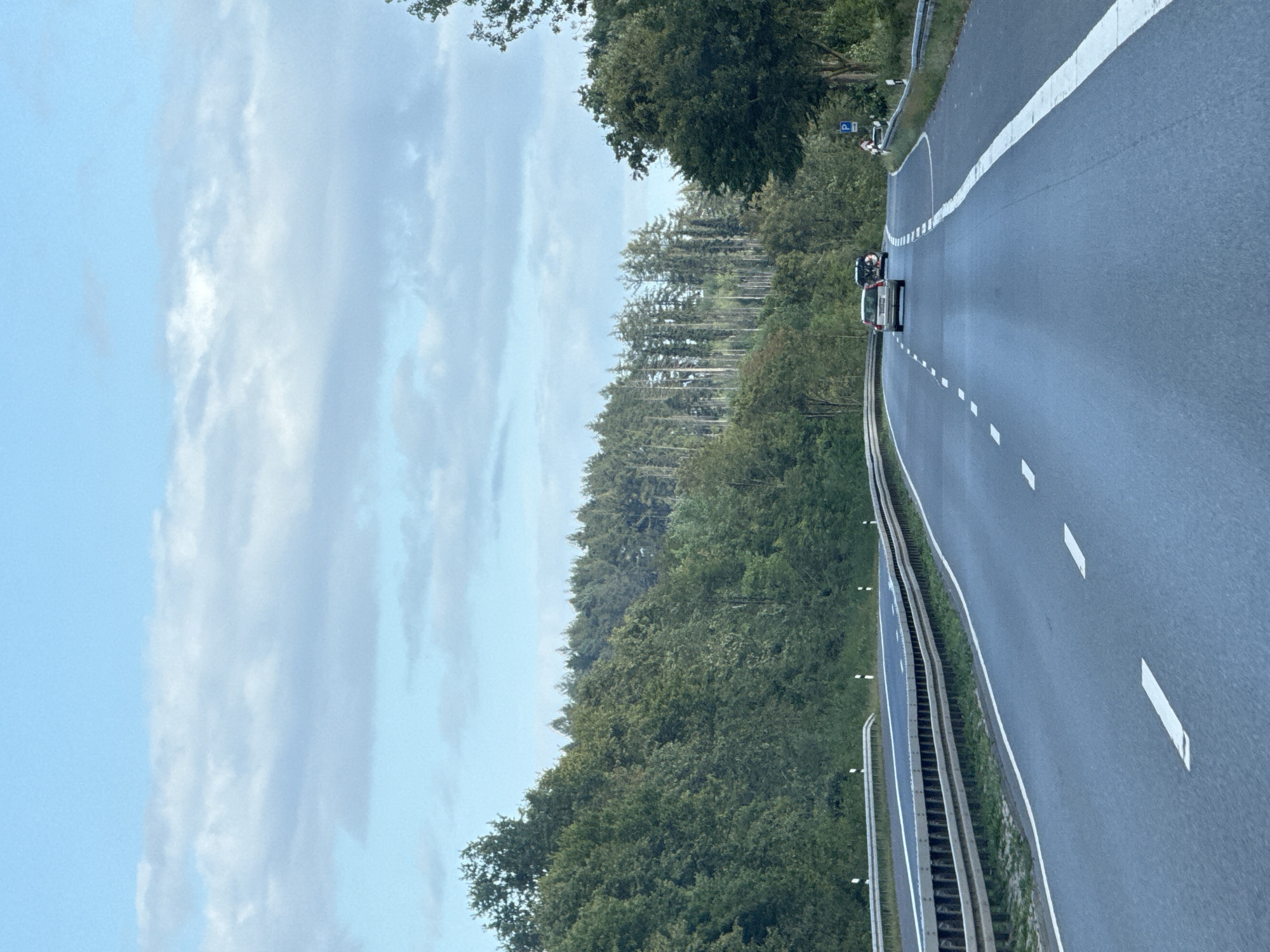 The image captures a view from a vehicle traveling on a highway in Herzberg, Germany. The highway is asphalt with white dashed lines indicating lanes and a solid white line marking the edge of the road. Guardrails line the sides of the highway. Dense green forests with a mix of deciduous and coniferous trees flank the road, with a stand of tall, slender coniferous trees in the background. The sky is partially cloudy with a light blue hue. Two cars are visible on the highway ahead, with a third vehicle further in the distance. A blue sign with a white 'P' symbol, indicating parking, is present on the right side of the road. The scene appears to be a typical highway environment in a rural or semi-rural area.