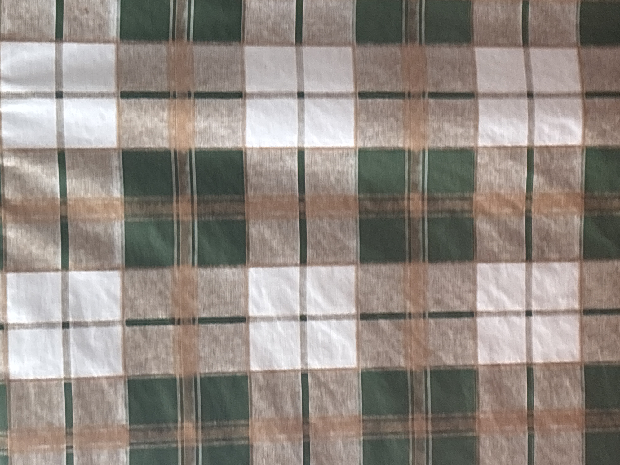 A close-up shot shows a plaid fabric with alternating stripes of green, brown, and white. The pattern consists of wide green stripes that are intersected by narrower brown and white stripes, forming squares. The lighting is even, and the image is in focus. There are no people or objects visible in the scene, and the background is simply the fabric itself. The location is listed as Unknown, Unknown.
