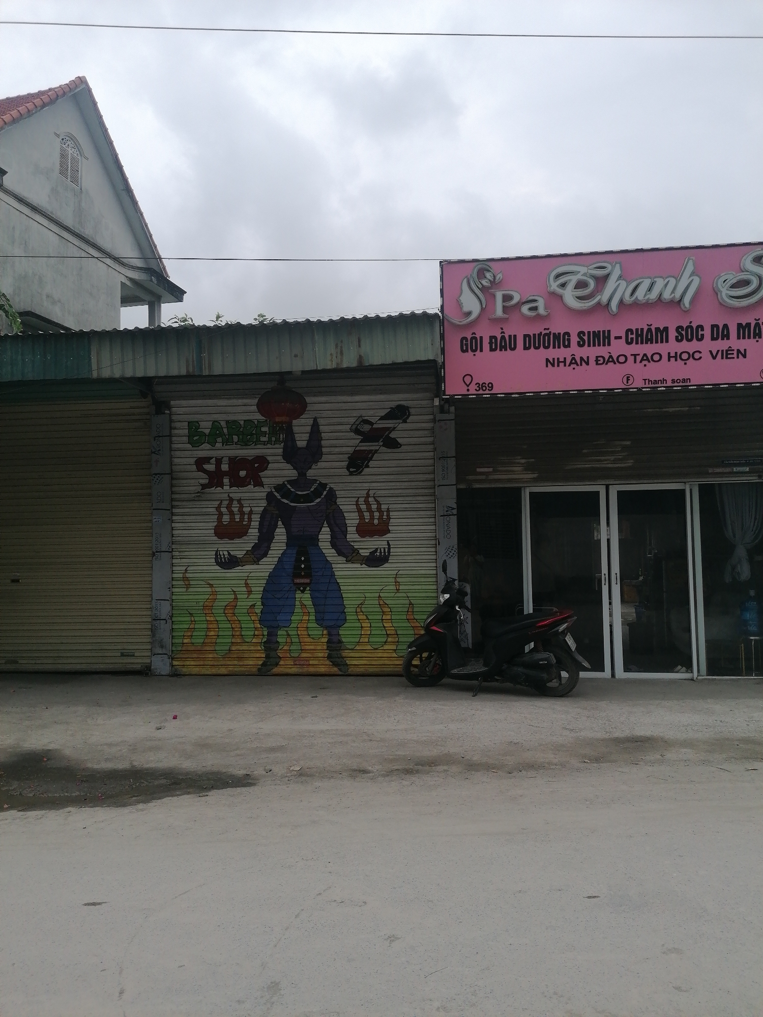 The image captures a street scene in Ha Tu Ward, Vietnam, featuring two storefronts side by side. On the left, a corrugated metal roll-up door is adorned with a colorful mural depicting the character Beerus from Dragon Ball Super, labeled as a "BARBER SHOP". To the right, a pink sign reads "Spa Thanh S", with Vietnamese text promoting hair and skincare services, and a business number "369" displayed. A black scooter is parked in front of the spa entrance, and a building with a pitched roof is visible in the background under a cloudy sky. The street surface appears to be concrete.