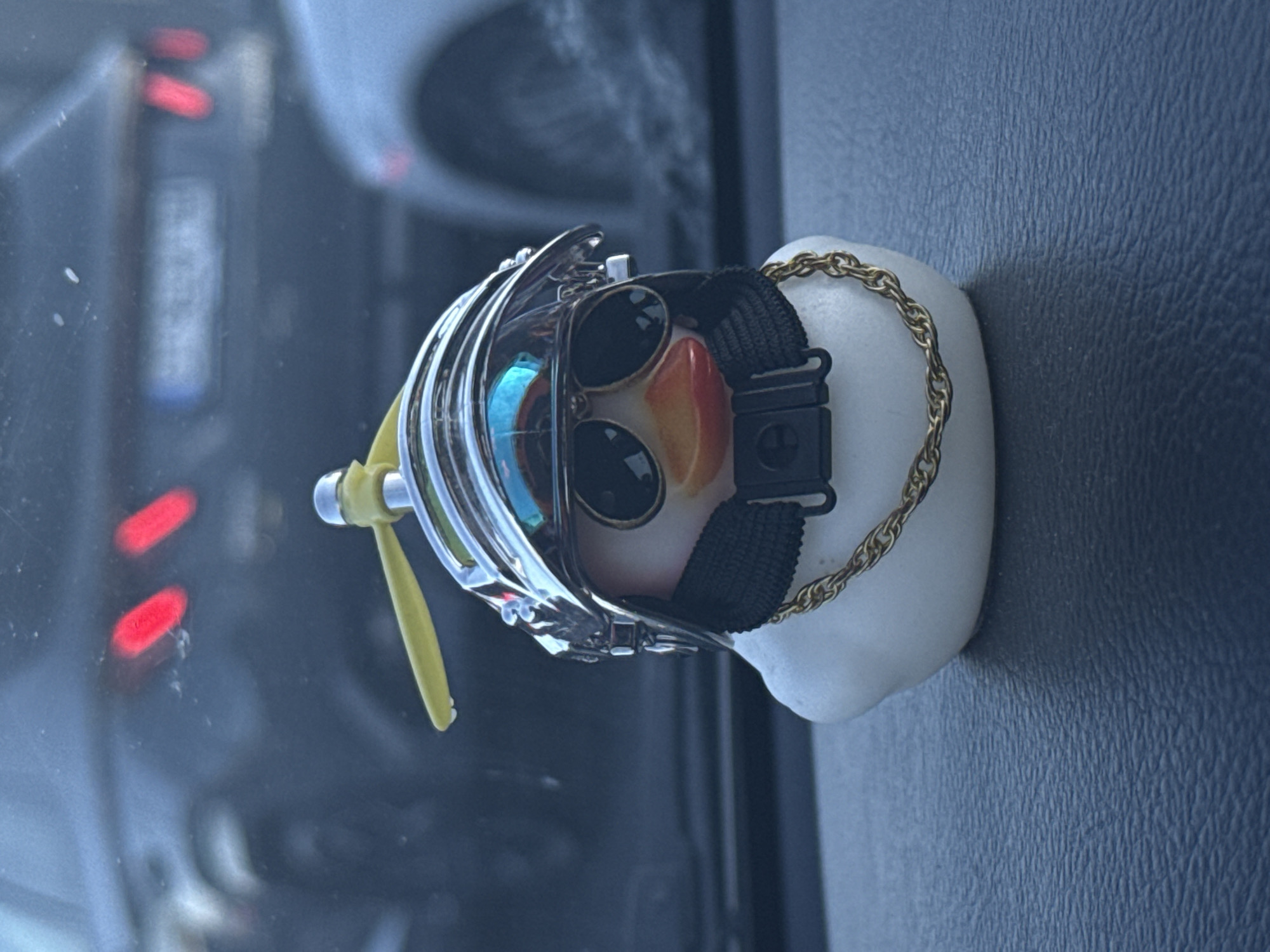 A close-up photograph depicts a small, white, toy duck adorned with sunglasses, a gold chain, and a silver aviator helmet with a yellow propeller. The object is positioned on a dark blue, textured surface, possibly the dashboard of a car, with a blurry reflection of a street scene visible through the windshield. The reflected scene hints at an urban environment, which might be Plovdiv, Bulgaria. Red tail lights are discernible in the background reflection.