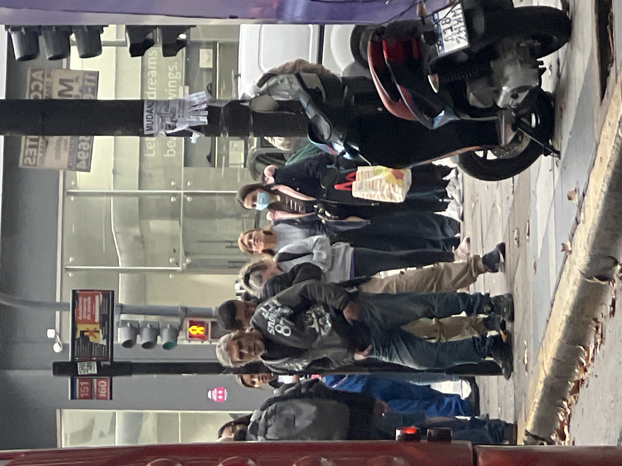 The image shows a group of pedestrians waiting on a sidewalk in Buenos Aires, Argentina. The pedestrians are arranged in a queue. Many of the pedestrians are facing forward. One woman is wearing a mask. A parked scooter is adjacent to the queue. Street signs "151" and "160" and a pedestrian crossing signal can be seen.
A sticker "Let Mudanz dreams be vings" is attached to a pole.