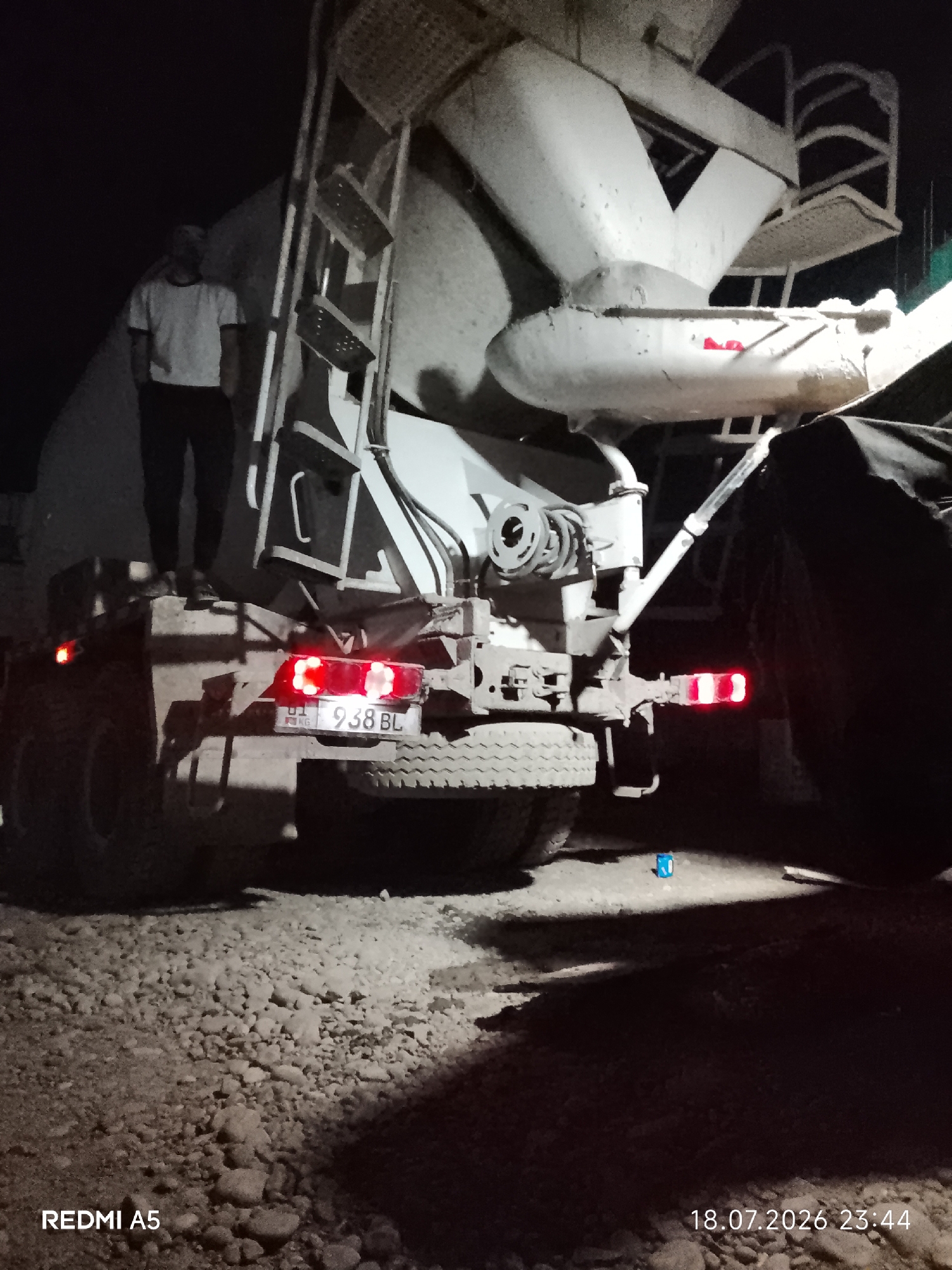 A concrete mixer truck is parked at night on a gravel surface in what appears to be an industrial or construction area, possibly in Tokmok City, Kyrgyzstan. A person wearing a white t-shirt and dark pants stands on the rear platform of the truck, looking away from the camera. The truck's rear lights are illuminated, and a license plate with the characters "938 BL" is visible. The ground is covered in small stones and dirt, and shadows are cast by the truck and the person. A small blue object is visible on the ground in the lower right portion of the image. The timestamp "18.07.2026 23:44" is displayed in the bottom right corner.