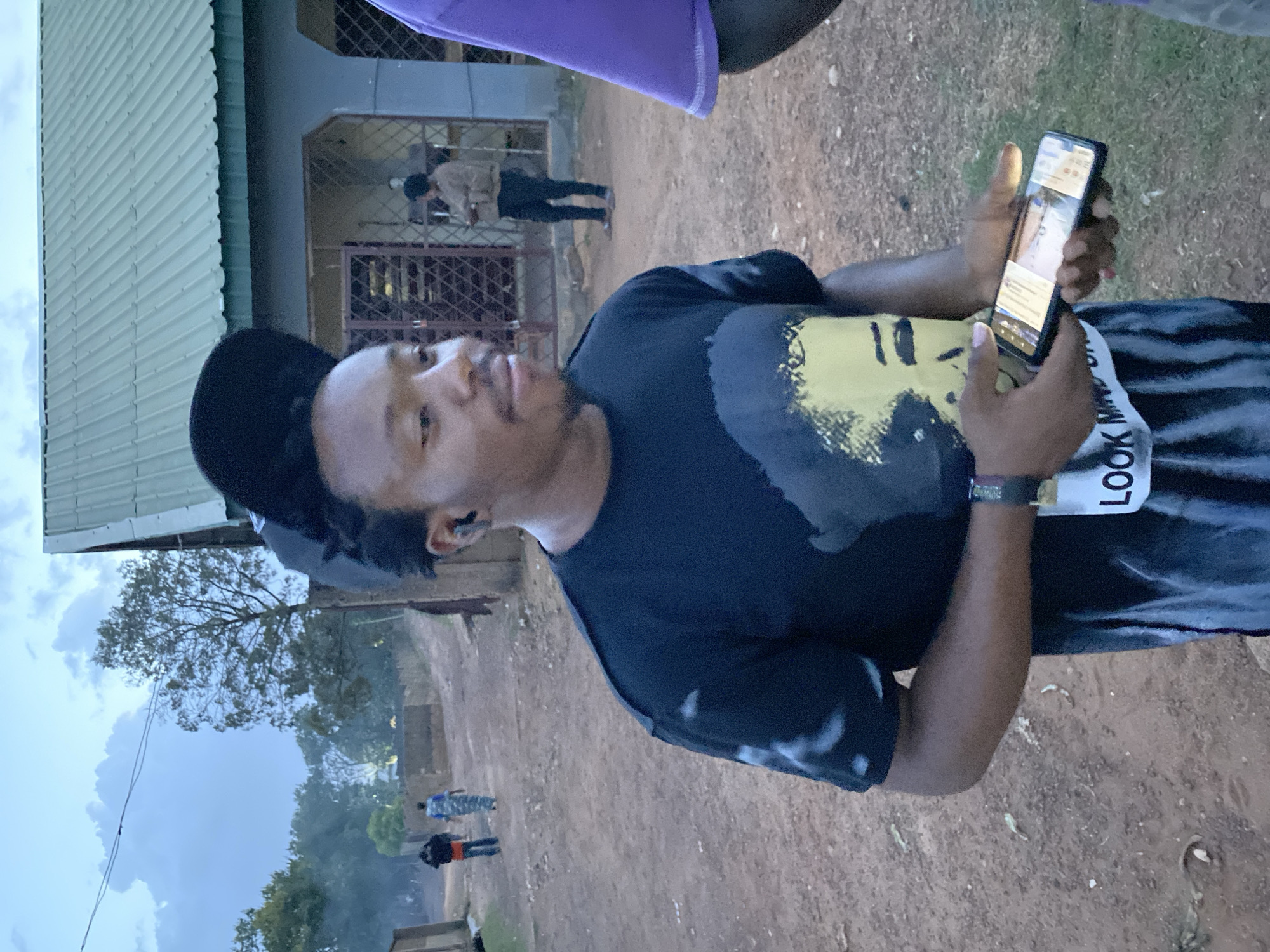 A person wearing a black t-shirt with a yellow graphic is looking at a mobile phone. The person has a black cap on their head and is wearing a black watch. The person also has a white race bib or sticker labeled with the text "LOOK MIND". The person is standing outside on a dirt path or road, with other people walking in the background. A building with a green roof and a gated entrance is also visible in the background. The photograph was taken in Bukuru, Nigeria, based on the geotag provided. The sky appears overcast.