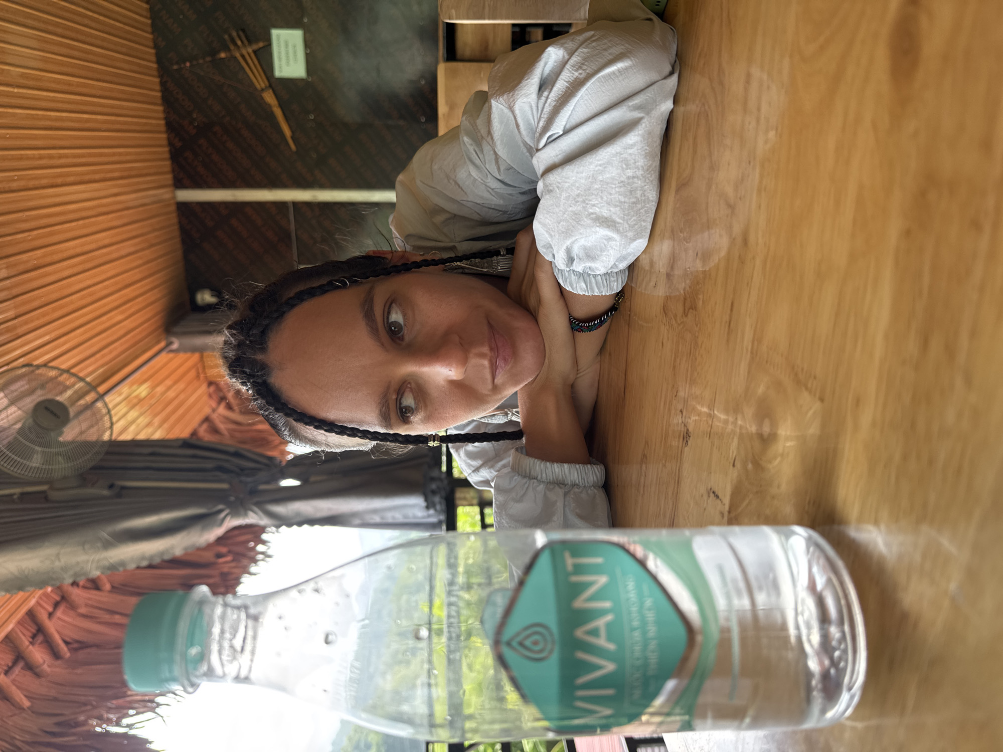 A woman with braided hair rests her head on her hands on a wooden table in Mậu Duệ Commune, Vietnam. A bottle of Vivant water sits in the foreground. A fan is visible in the background, along with a thatched roof. The lighting is bright.
