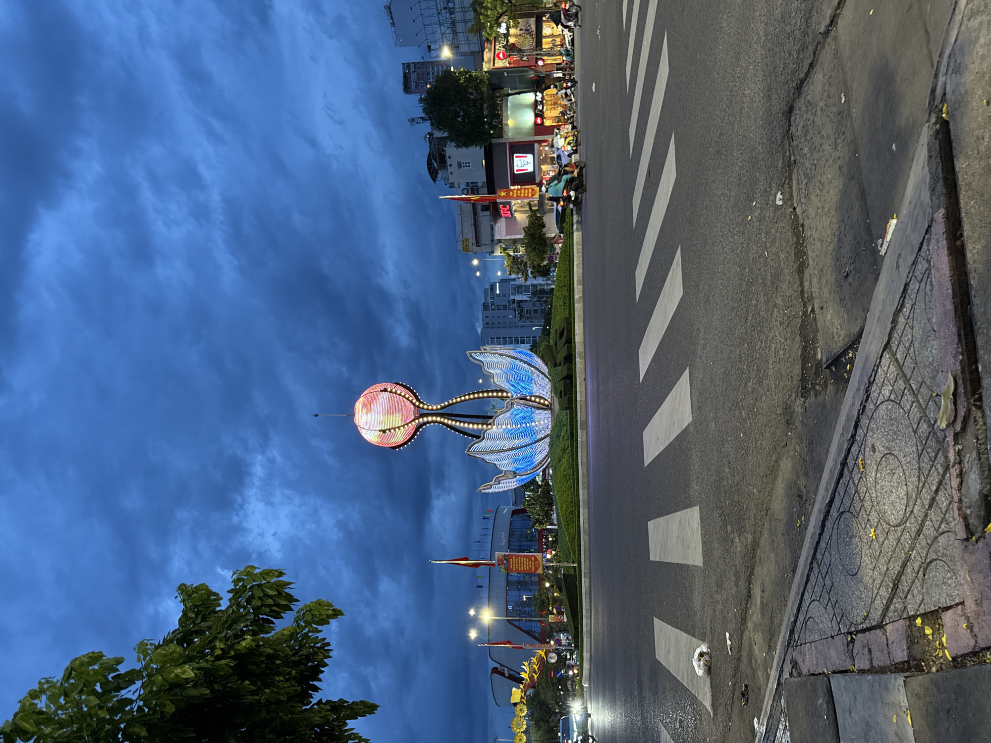 The image depicts a street scene at dusk in Nha Trang, Vietnam, focusing on a large, illuminated lotus-shaped sculpture in the center of a roundabout. The sculpture features a glowing orange and yellow top and a blue, petal-like base. The sky is a deep, cloudy blue, suggesting an approaching storm or recent rainfall. A tree with green leaves is visible in the upper left corner. The roundabout has a paved surface with greenery. Multiple traffic lights and streetlights illuminate the surroundings. Buildings, including businesses like KFC and Pizza Hut, line the street in the background. Several motorcycles and a car are on the road, with some riders visible. Pedestrian crossings with white stripes are present on the asphalt. The foreground shows a paved sidewalk with decorative patterns and some scattered yellow leaves. There is some litter on the road and sidewalk.
