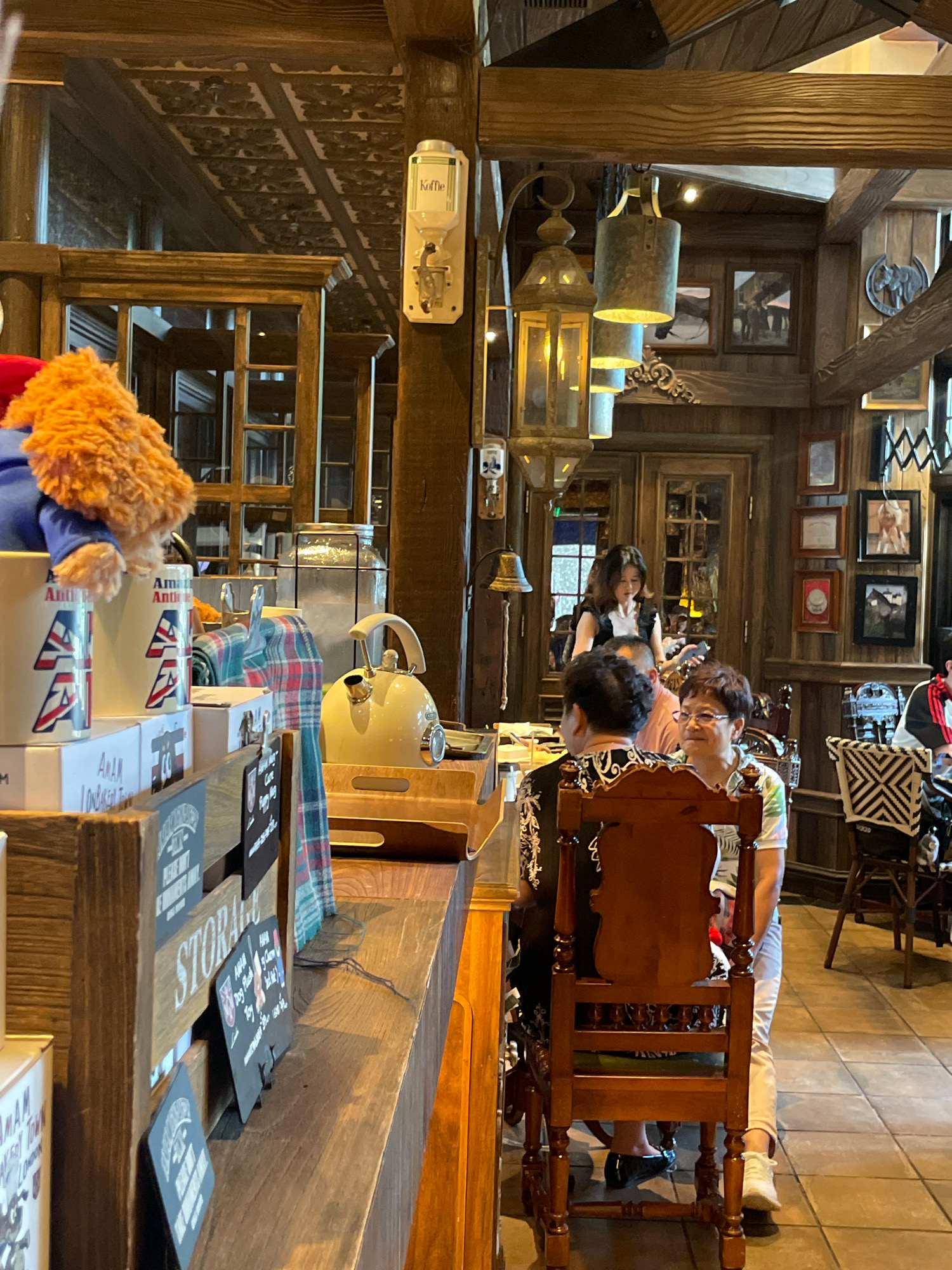 This is a medium-shot, eye-level, interior photograph of a cozy, rustic-themed cafe, possibly in Shanghai, China, given the context of the query. The cafe features wooden paneling on the walls and ceiling, with exposed beams and decorative metalwork. A stuffed bear wearing a blue coat and red hat is placed on a counter in the foreground, next to two mugs with British flag designs. To the right of the bear, a vintage-style cream-colored kettle sits on a wooden board, accompanied by a red and blue checkered cloth.
A counter extends across the bottom of the frame, displaying various retail items, including boxes and small signs. One sign clearly reads "STORAGE". In the middle ground, several patrons are seated at tables. A woman with dark hair is standing behind a counter, interacting with a customer. Two women are seated at a table on the right side of the frame, one facing the camera, wearing glasses and a patterned shirt, and another facing away.
The overall atmosphere suggests a comfortable and inviting establishment, possibly specializing in British-themed merchandise or food. The lighting is warm and subdued, enhancing the rustic ambiance.