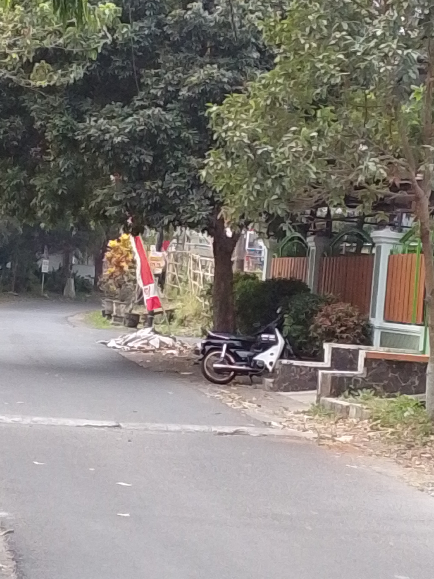 A motorcycle is parked on the side of a paved road in front of a residential property in Malang, Indonesia. The motorcycle is white and black with a silver exhaust pipe. The property features a fence with wooden slats and decorative pillars. A red and white flag, likely the Indonesian flag, is planted near the entrance to the property. Trees and foliage surround the scene, with some leaves scattered on the ground. The road appears to be in moderate condition, with some cracks and debris.