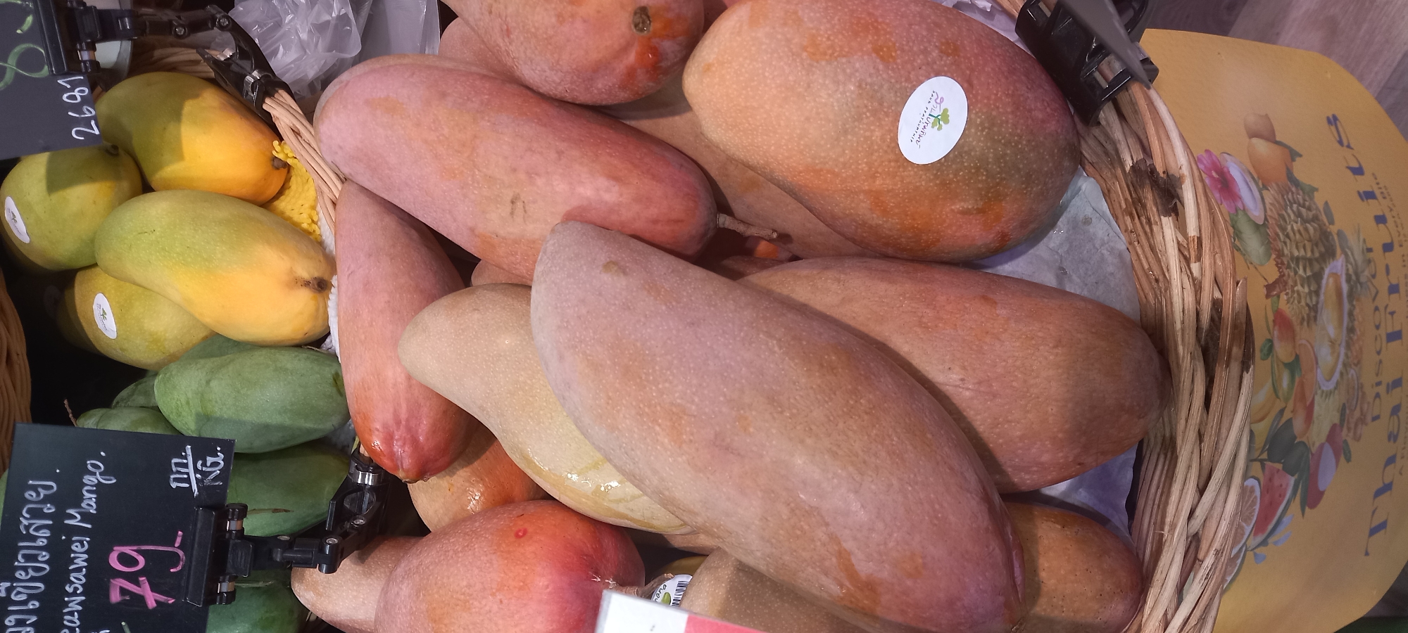 A basket of mangoes, including green, yellow, and pink varieties, sits on a produce display. A price tag reads "เขียวเสวย" (Keaw Sawei Mango) with a price of 79. The address "Moo 1" and the words "SPUN PRATHUMTHIP" indicate this location is in Thailand. A sticker on one mango reads "สวนปทุมทิพย์" (Suan Prathumthip), which means "Prathumthip Garden." The display also includes a sign that says "Discover Thai Fruits."