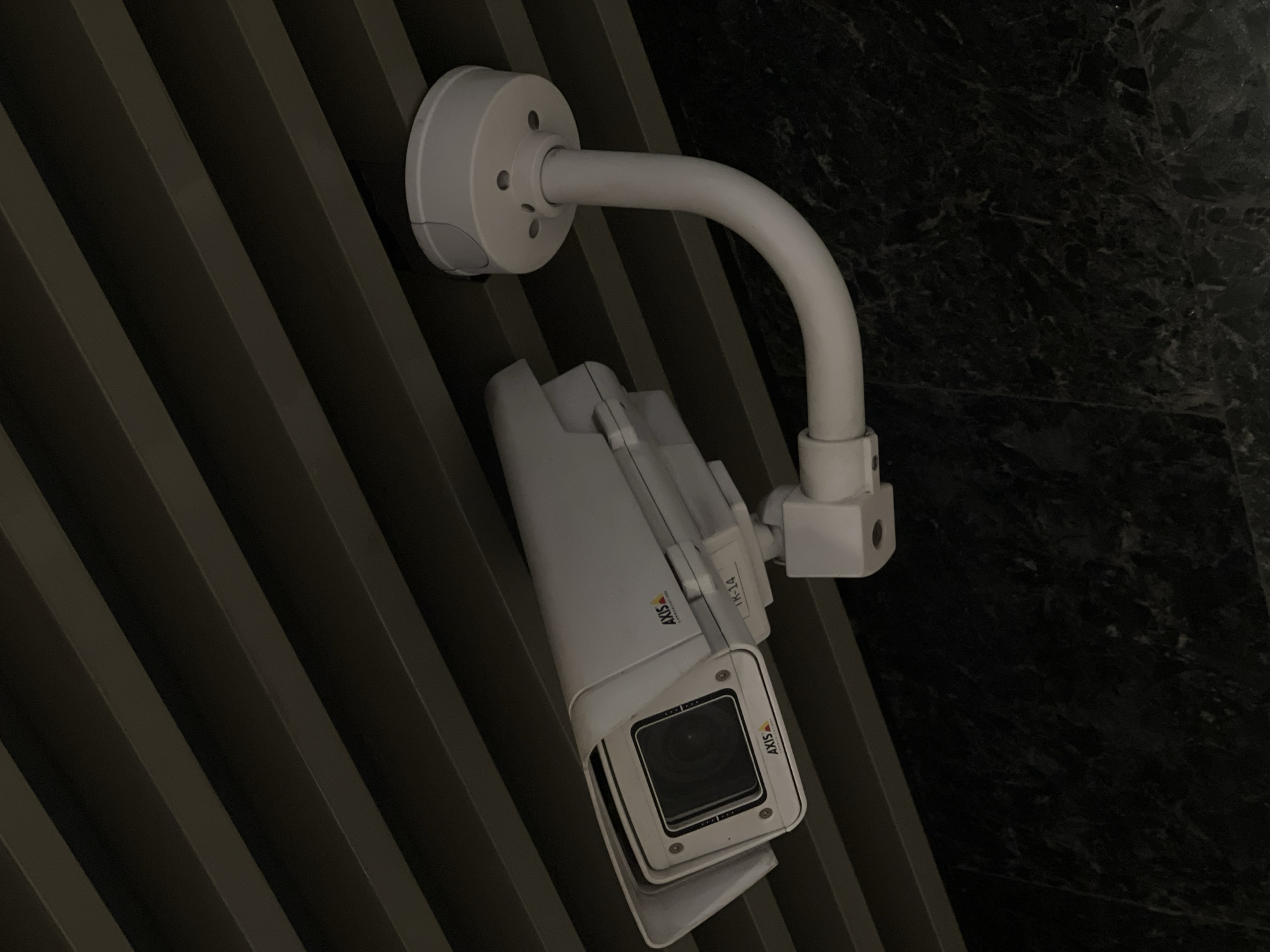 A white AXIS brand security camera is mounted on a wall. The wall is textured with a series of gray diagonal stripes. The camera is attached to the wall with a white, rounded mount and a curved arm. The camera is angled downwards. To the right of the wall is a dark, mottled surface that appears to be stone or marble. There is no evidence to indicate it is in Moscow, Russia.