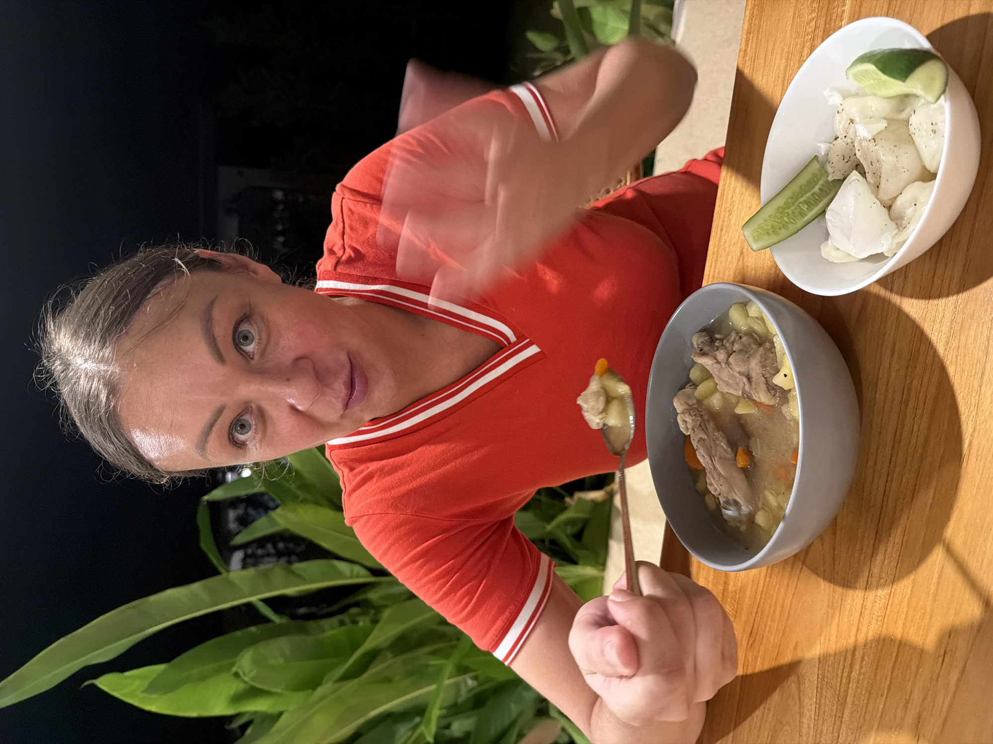 A woman with shoulder-length brown hair and light blue eyes is seated at a wooden table outdoors at night, with lush green plants in the background. She is wearing a red t-shirt with white trim. The woman is holding a spoon with food in her right hand and is looking at the camera with a surprised expression. A gray bowl filled with soup containing meat, potatoes, and carrots is in front of her, and she is in the process of bringing a spoonful of soup to her mouth. To the right of the soup bowl is a smaller white bowl containing chunks of white food, likely fish or chicken, garnished with slices of cucumber and lime. The scene is illuminated by artificial light, casting shadows on the wooden table. The location is identified as Koh Phangan, Thailand, likely a restaurant or outdoor dining area.