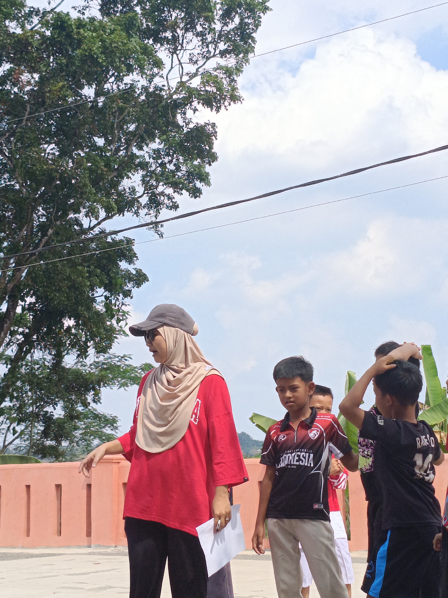 A group of children and an adult are gathered outdoors on a sunny day, possibly in Kadununggal, Indonesia. A woman wearing a red shirt, beige hijab, and a baseball cap stands on the left, holding a piece of paper. Several boys are on the right, one wearing a black and red shirt with "INDONESIA" printed on it, and another in a black shirt with the number "14". They appear to be interacting with each other. The background shows a pink wall, a large tree with green leaves, and a cloudy sky with power lines.