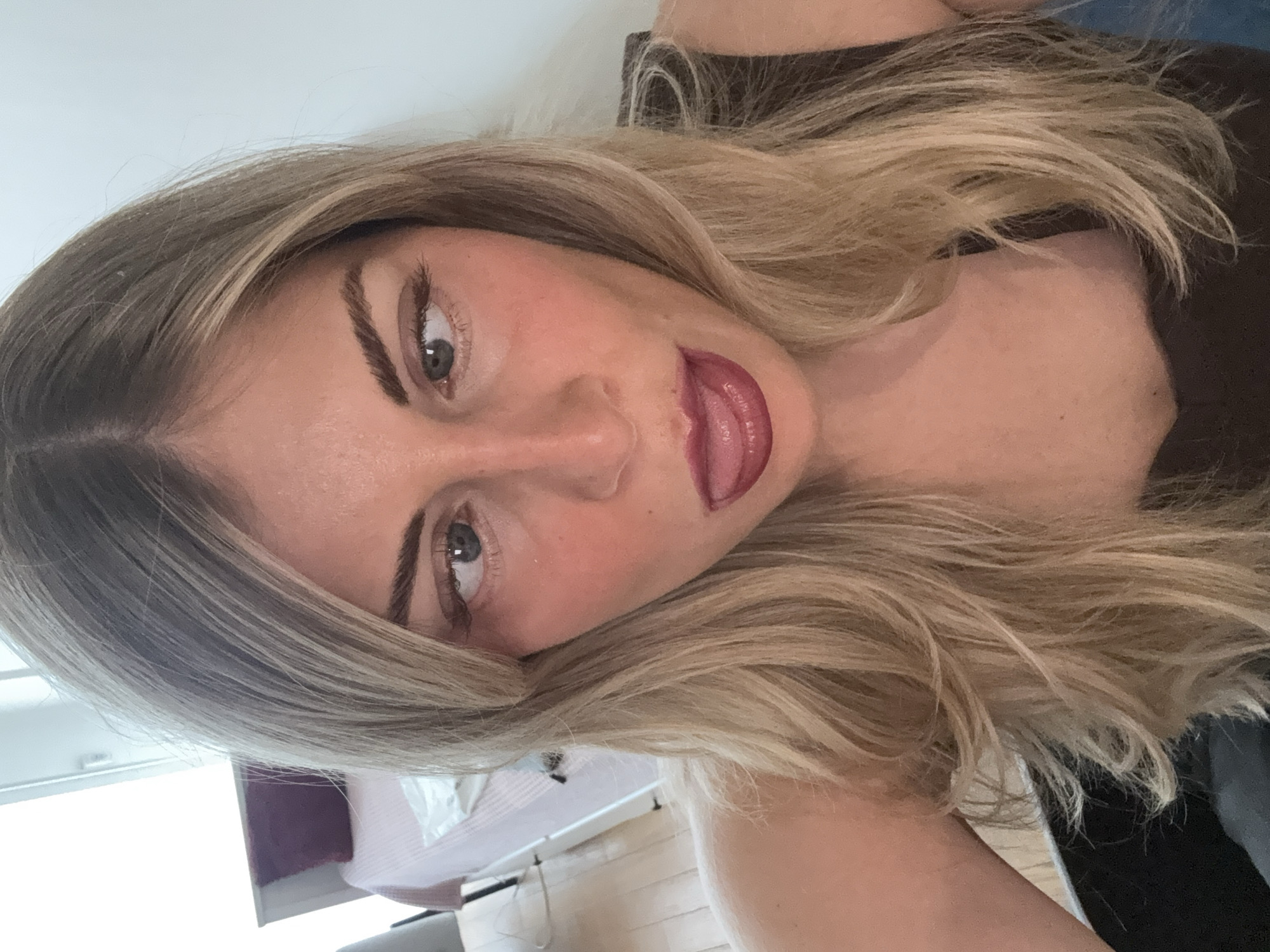 The media file presents a close-up portrait of a young woman, captured in a selfie style with a significant counter-clockwise tilt, orienting her head at an angle. She has fair skin, prominent and well-defined dark eyebrows, and light-colored eyes (likely green or hazel) that gaze slightly towards the right. Her full lips are adorned with a dark pink or berry-toned lipstick, and her tongue is playfully extended, touching her upper lip. Her long, wavy hair is a mix of blonde and darker roots or highlights, cascading around her face and shoulders.
She is wearing a dark, likely black or brown, top or dress. The background is softly blurred, indicating an indoor setting with natural, bright daytime lighting. Visible elements in the background include a light-colored wall, and hints of domestic furniture such as a bed with a light-colored duvet and a purple headboard or cushion, and a wooden floor. Her expression is playful and engaging. There is no visible text in the image. This media is contextualized as being from Paris, France.