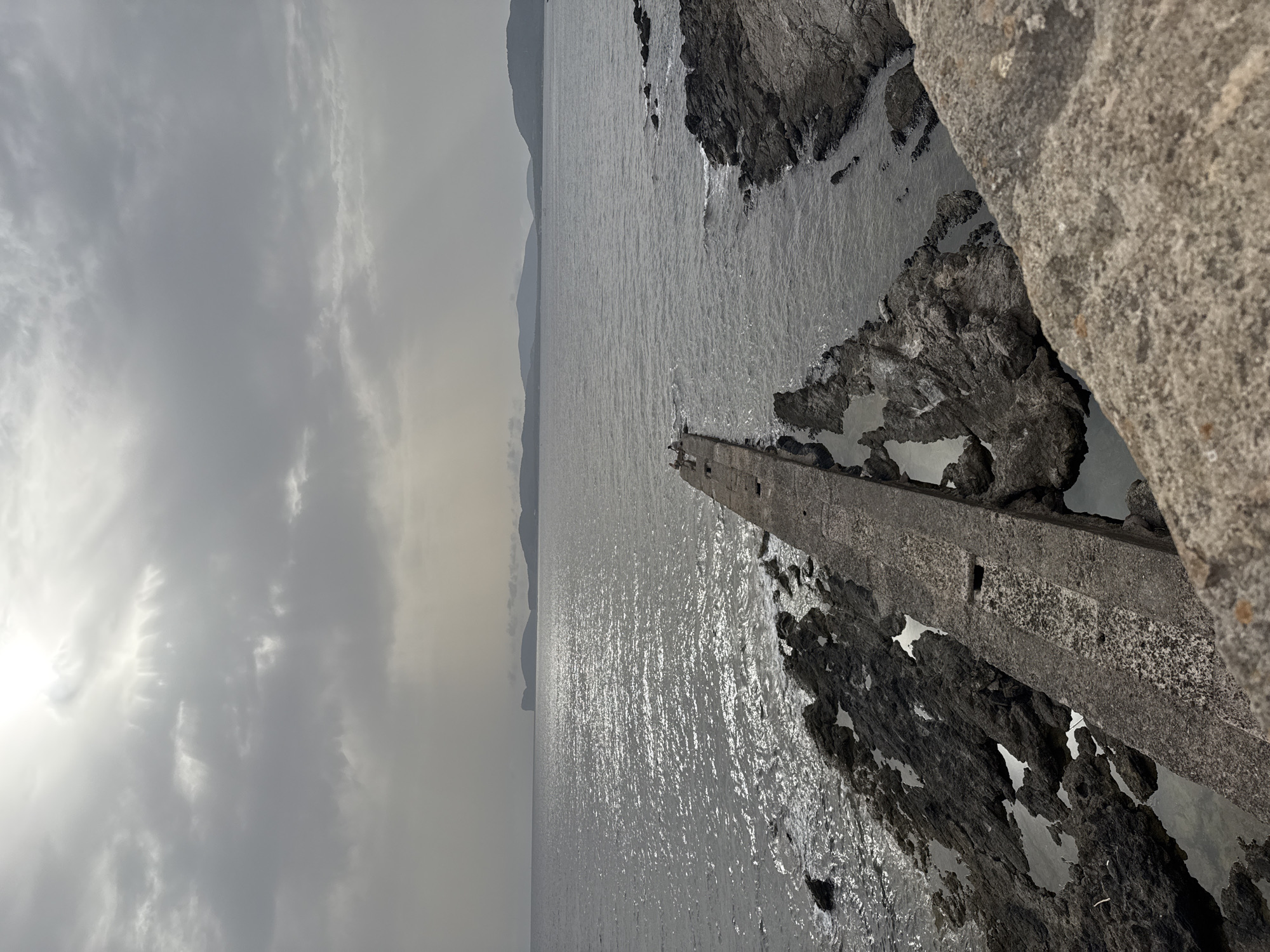The image depicts a dramatic seascape with a concrete structure extending into the water. The sky is overcast with a bright, diffused light source on the left, suggesting it might be late afternoon or early morning. The sea is choppy, with sunlight glinting off the surface, and it meets a rugged coastline of dark rocks. The concrete structure appears to be a pier or a breakwater, showing signs of wear and tear. In the distance, hazy mountains form a silhouetted horizon. The overall mood is somewhat somber and imposing, highlighting the raw power of nature against man-made elements. The location context is Alghero, Italy.