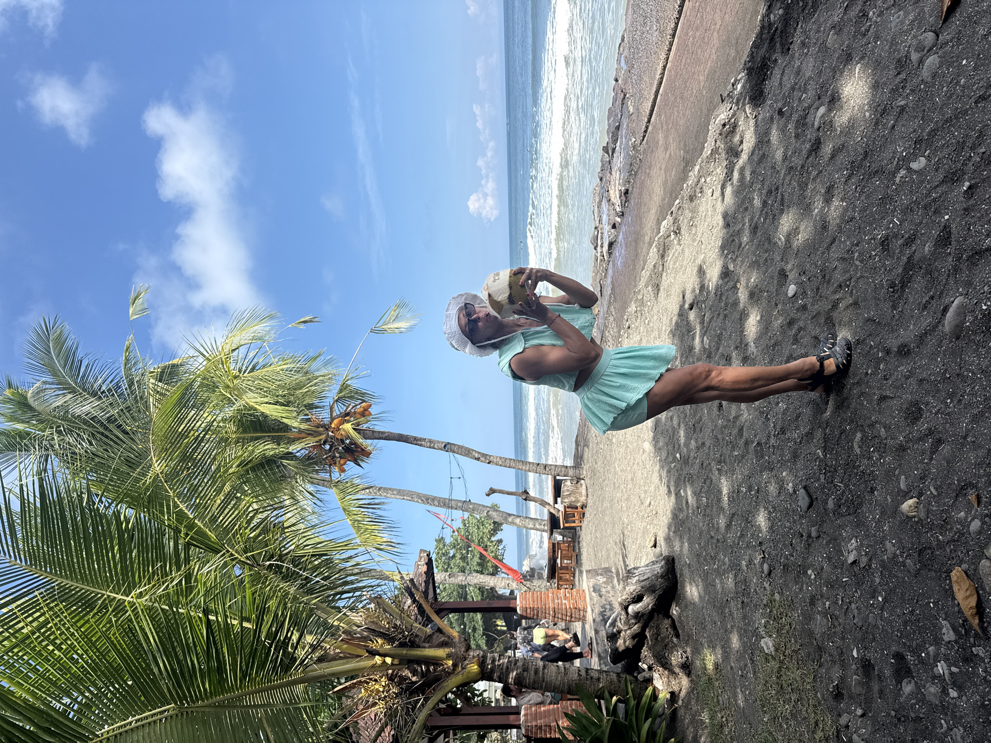 A woman stands on a beach in Unknown, Unknown, holding a coconut to her mouth. She wears a light green dress, white hat, and black sandals. The beach has black sand. The ocean is in the background, with waves visible. Behind the woman is a palm tree and part of a building. The sky is blue with scattered clouds.