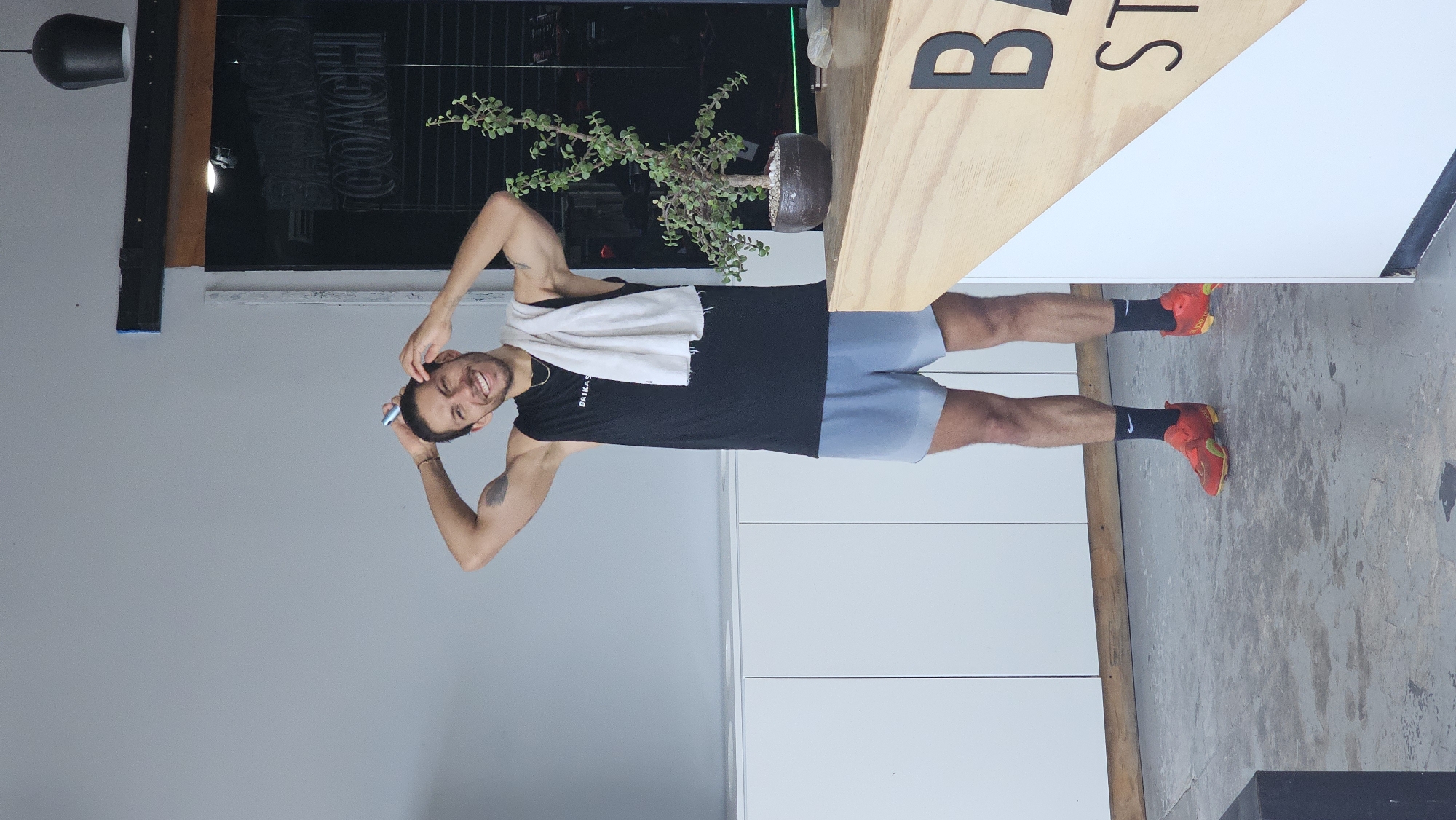 The image, taken in Puebla, Mexico, features a smiling young man posing indoors, likely within a gym or fitness studio. He has light brown skin, dark hair, and a light beard, and is wearing a black sleeveless shirt with "BAIKAS" visible on it, light blue shorts, and bright red sneakers with black socks. A white towel is draped around his neck. He smiles broadly at the camera, holding a small blue object (possibly a phone or vape pen) near his head with his right hand. A tattoo is visible on his left bicep.
The background includes a grey wall on the left, with a black board or surface behind him that has the word "COACH" written on it in a stylized, backward manner. A dark light fixture hangs from the ceiling. To his right, a light wooden partition or counter displays large black letters, partially visible as "B ST". A potted plant with small green leaves sits on a surface behind this wooden structure. The floor is grey, concrete, and textured. The indoor setting is brightly lit, suggesting it is daytime. The man's cheerful expression contributes to an overall positive and energetic mood.