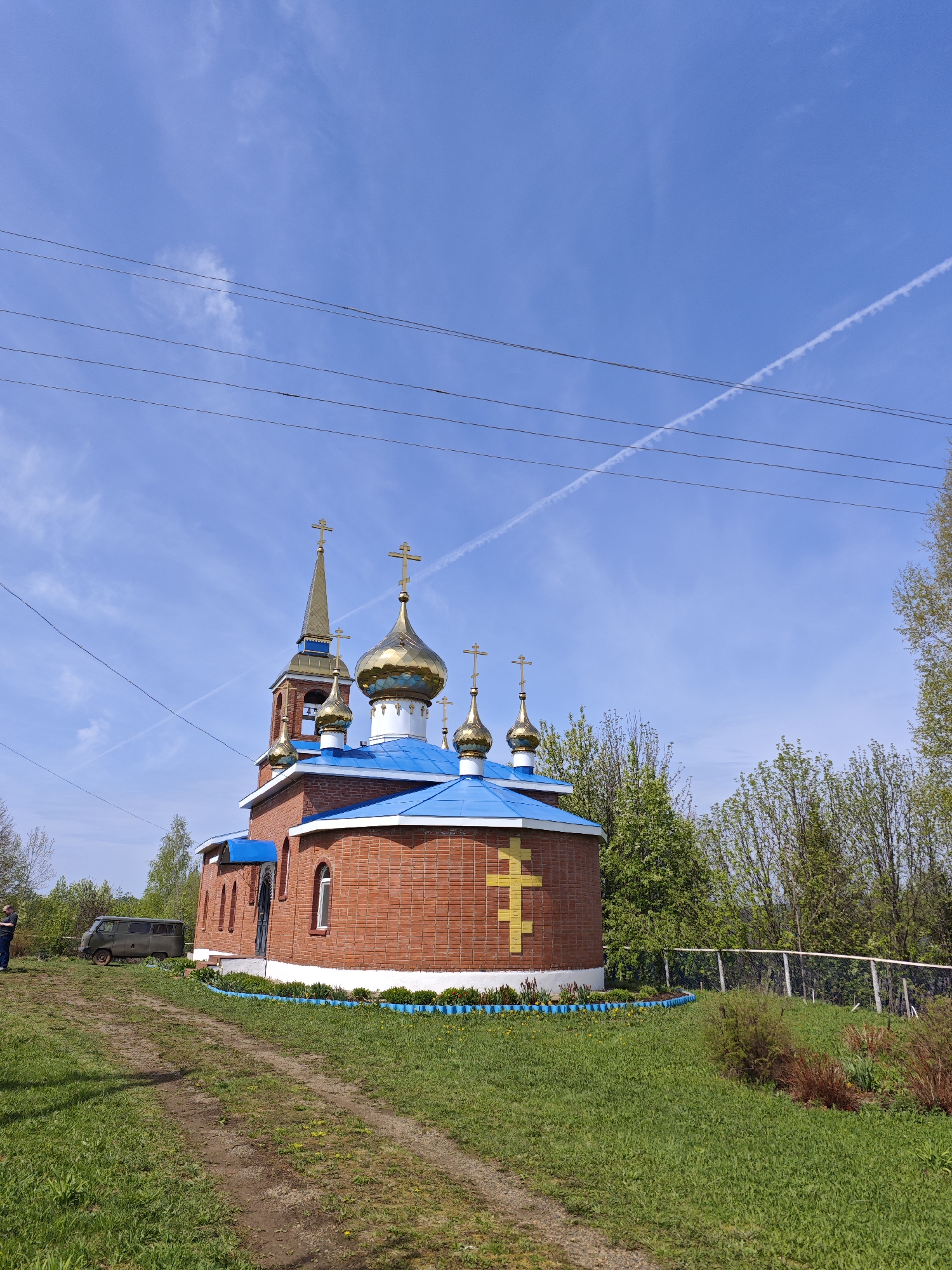 The image depicts a brick Russian Orthodox church with a blue roof and multiple golden domes, located in Большая Уса, Russia. The church is surrounded by a green lawn and a small decorative fence. A dirt path leads up to the church. In the background, there are trees and a white fence. Power lines stretch across the upper portion of the frame. A faint contrail cuts across the blue sky. A person and a dark-colored van are faintly visible on the left side of the image. The day is sunny.