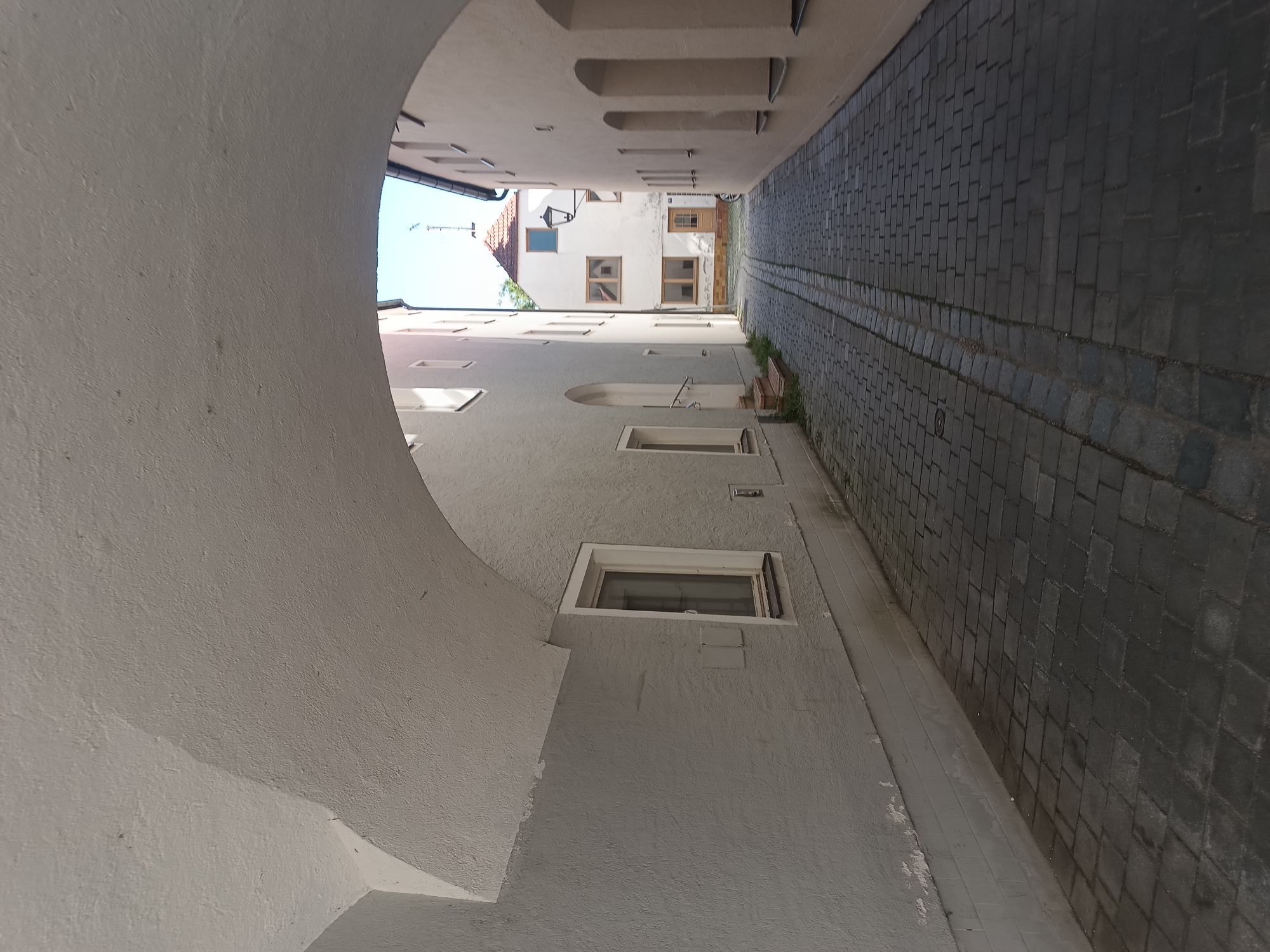 The image shows a narrow, cobblestone alleyway in the historic Old Town of Neuötting, Germany, as viewed from beneath an arched, stone passageway. The passageway has a rough, textured ceiling. To the left, a light gray building features a narrow window and a small, square metal fixture on the wall. Further down the alley, on the right, a building with cream-colored stucco walls has several arched recesses and windows. At the end of the alley, a white building with a red-tiled roof is visible, with windows and a wooden door. A vintage street lamp is mounted on the white building. The sky is clear and blue. The ground is paved with dark gray cobblestones, with a slight incline leading away from the viewer. There are no people visible in the scene.
