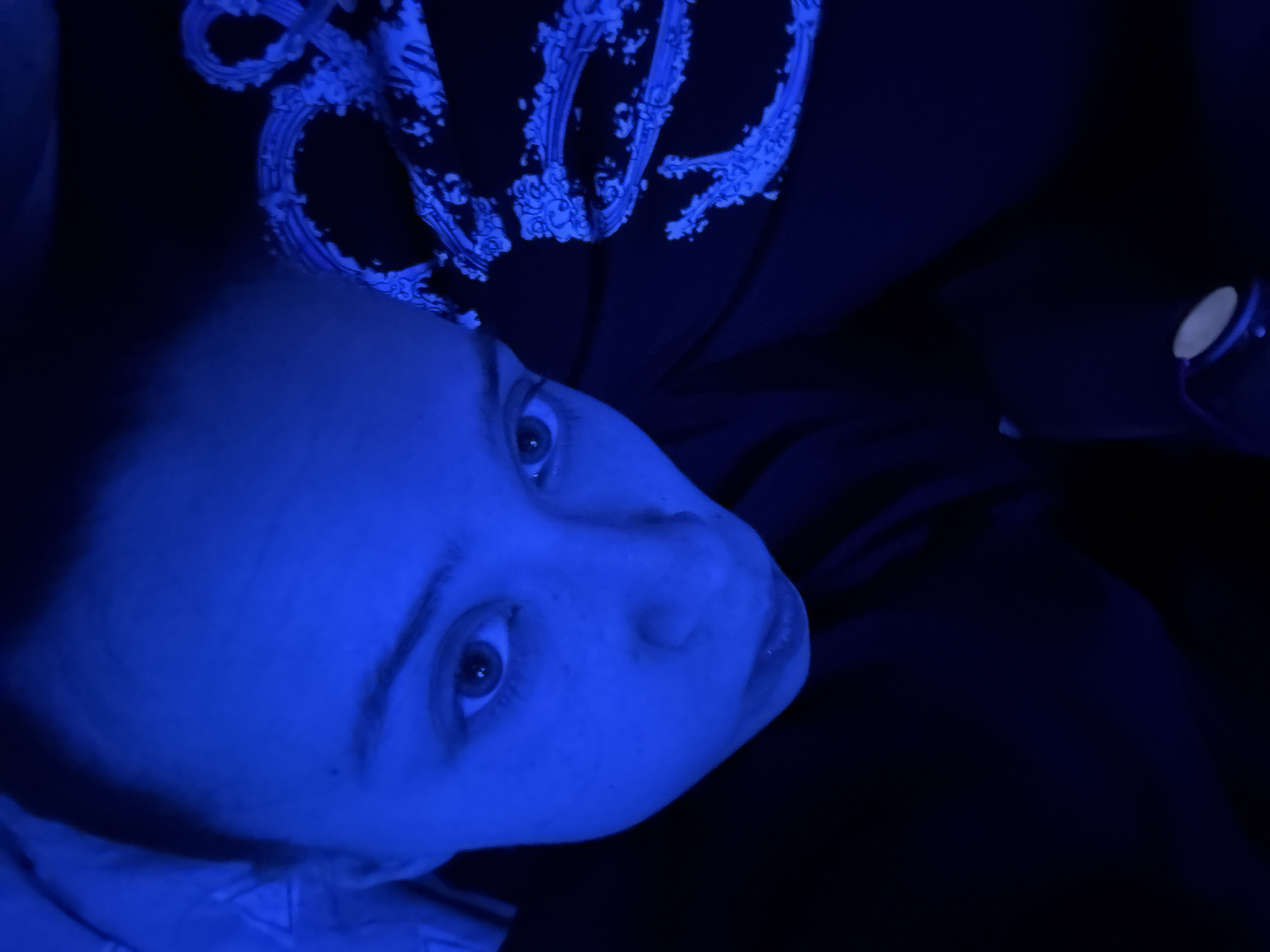 A close-up, angled shot of a person's face under a blue light. The person has light skin, and their eyes are visible and directed upwards. They are wearing a dark garment with a white design on the upper portion of the frame. The background is dark and mostly out of focus, with a round object visible on the right side. There is no definitive information about the location being in Прикамский, Russia.