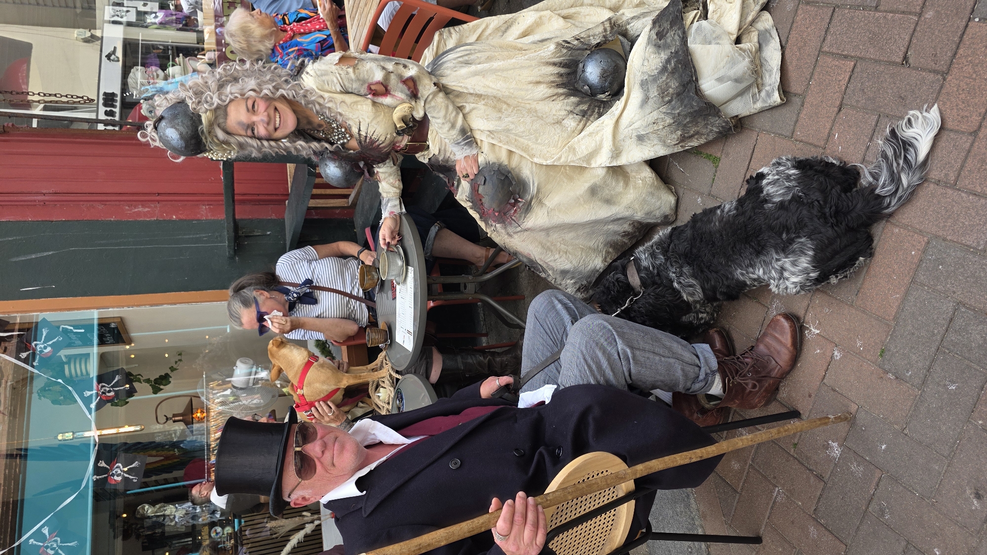 The image depicts an outdoor scene in St Leonards, United Kingdom, likely outside a shop or cafe. In the foreground, a man wearing a black top hat, sunglasses, and a dark coat with a maroon vest sits on a chair, holding a walking stick. To his right, a tan dog stands on a table. Further back, a woman in a striped shirt dabs her nose with a napkin. To the right, a woman with elaborate, curly gray hair and a distressed, cream-colored gown, adorned with dark, spherical objects, smiles at the camera. She is seated and a black and white dog lies at her feet. The environment includes a paved ground, a red-fronted building, and a shop window displaying pirate-themed decorations.