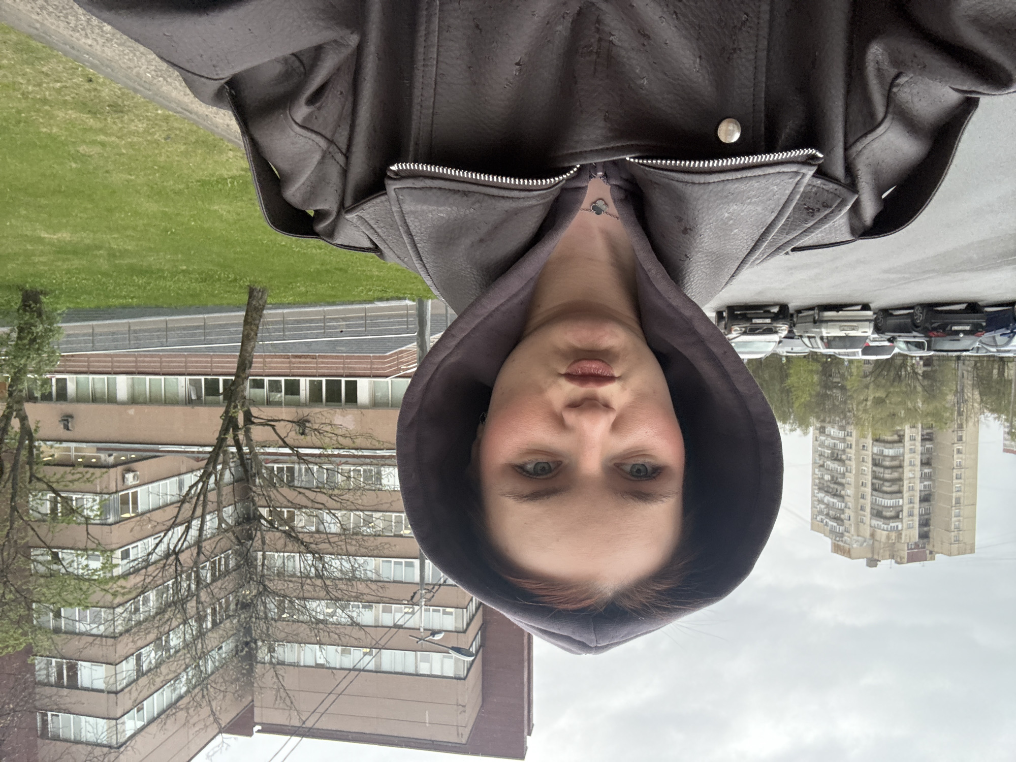 A woman wearing a brown leather jacket with the hood up is posing for a selfie. Behind her is a cityscape with buildings, trees, and parked cars. Based on the architecture of the buildings, the scene is likely located in Saint Petersburg, Russia. The sky is overcast.