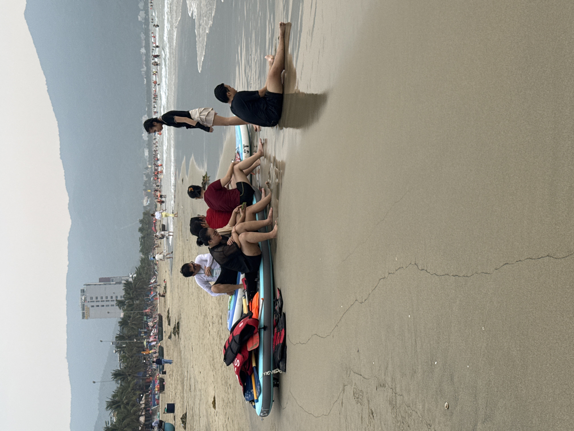 The image depicts a group of people sitting on a large paddle board on a beach. There are approximately seven people, with some sitting and some standing. They are near the shoreline, with the ocean visible in the background. In the foreground, there is a pile of life jackets and paddles on the paddle board. The beach is sandy with some moisture. In the background, buildings, palm trees, and mountains are visible, indicating the location is in the city of Đà Nẵng, Vietnam. Many other people are on the beach in the background.