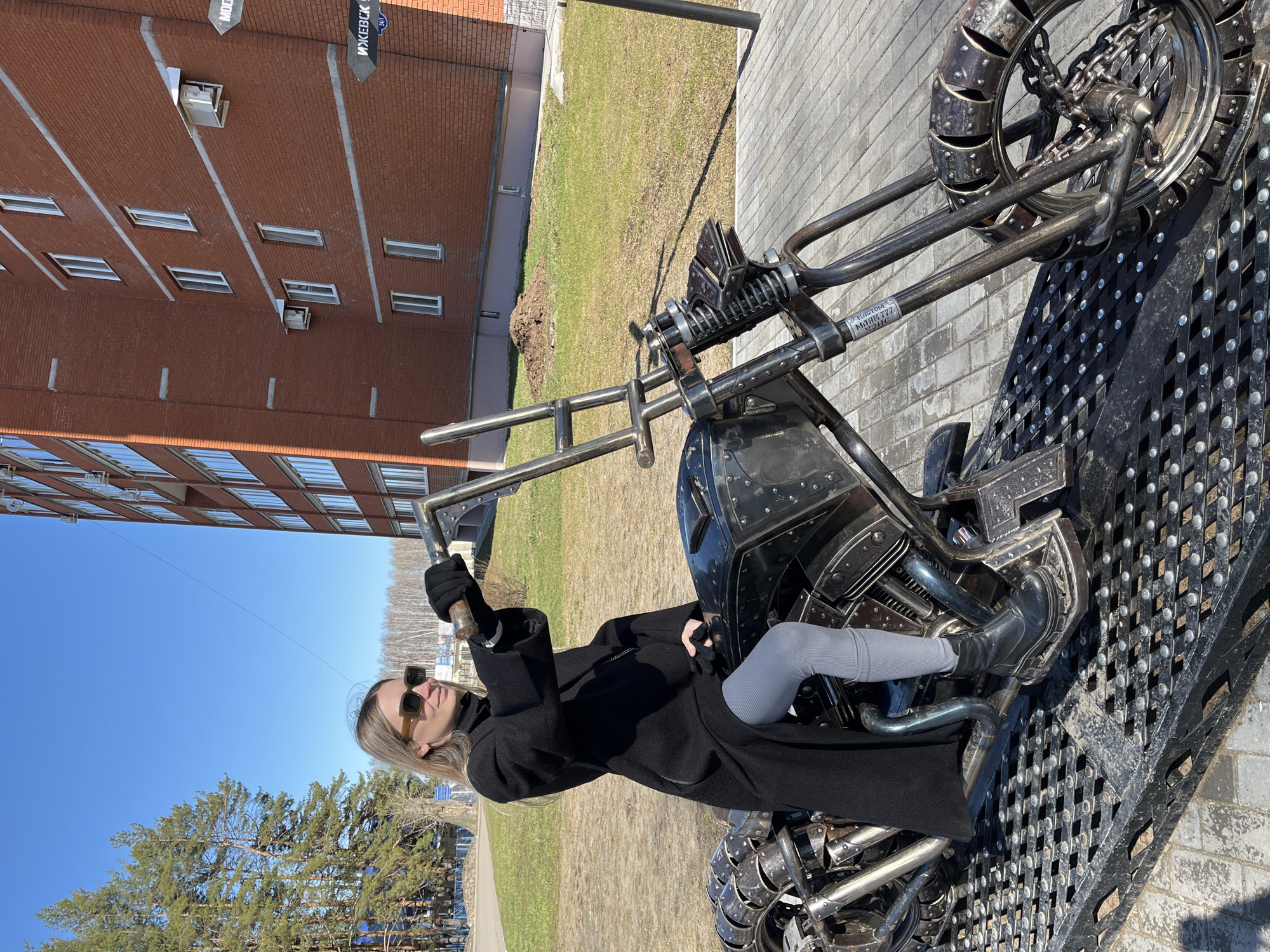 A woman with blonde hair and sunglasses is sitting on a metal motorcycle sculpture in the city of Chaikovsky, Russia. She is wearing a long black coat, gray leggings, and black boots. The sculpture is placed on a metal grid platform and features intricate metalwork, including chains and rivets. In the background, a multi-story brick building with multiple windows and a blue sky are visible. There is also a grassy area and some trees in the background.