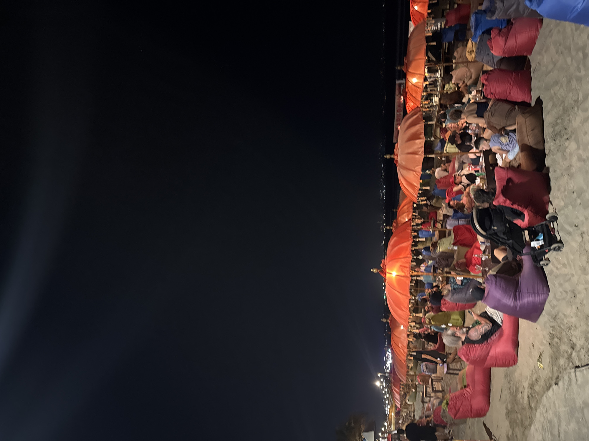 The image depicts a lively beach scene in Kuta, Indonesia, at night. Numerous orange umbrellas, illuminated from within, provide shade and ambiance over seating areas scattered across the sand. People are seen relaxing on beanbag chairs and at tables, engaged in conversations and enjoying the evening. The background reveals the distant city lights and a dark, vast sky. The overall atmosphere suggests a popular beachfront establishment, likely a bar or restaurant, catering to beachgoers seeking a nighttime experience.