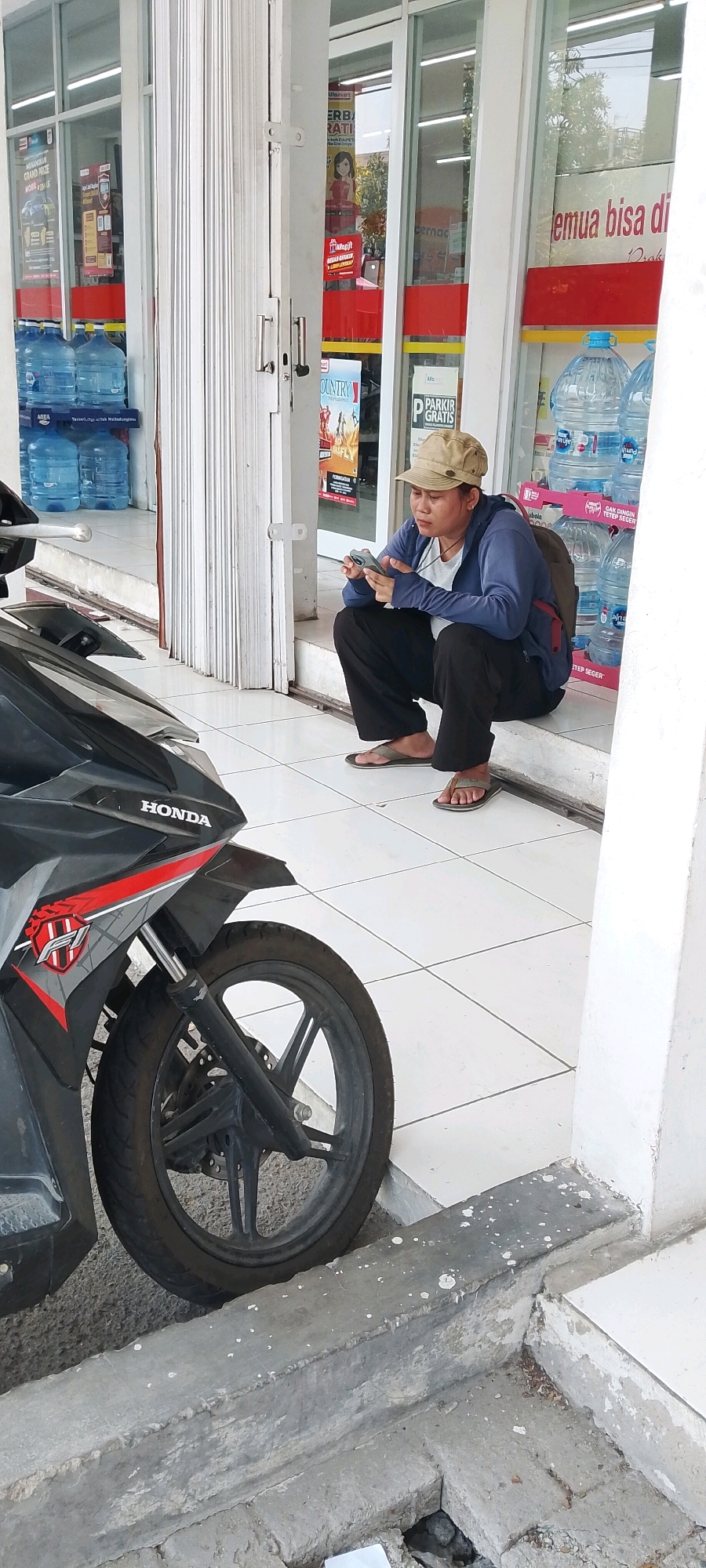 In Satriamekar, Indonesia, a person wearing a light brown cap, a blue jacket, and dark pants crouches on a tiled step outside a store. They are looking at and holding a smartphone. A black and red motorcycle is parked to the left of the frame, partially obscuring the lower left corner. Behind the person, stacked bottles of water are visible through the store's glass doors and windows, along with various promotional signs. The store appears to be a convenience store, suggested by the water bottles and signage. The area is outdoors, with a concrete curb in the foreground.