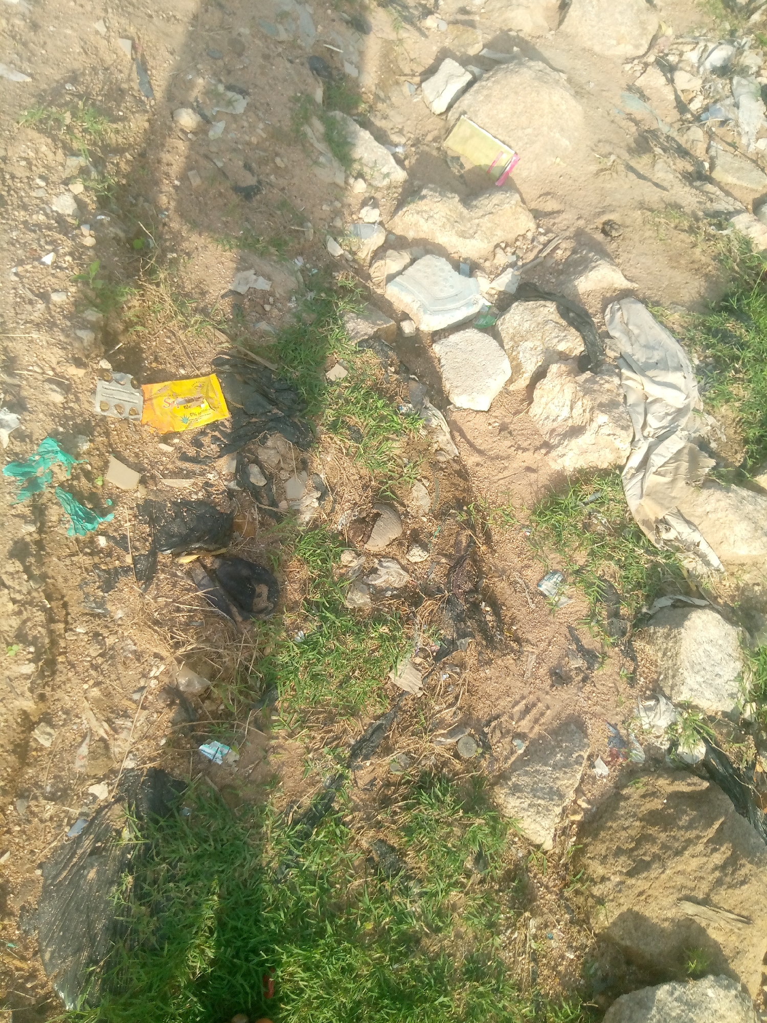 This image depicts a patch of uneven, sandy ground with scattered rocks and sparse green vegetation, possibly in Jauro Yinu, Nigeria. The ground is littered with various debris, including what appears to be torn plastic bags (some green, some black), crumpled paper or packaging, and possibly a small, light-colored cardboard box. A yellow wrapper with some text and a green leaf graphic is visible among the debris. Shadows suggest an overhead light source, likely the sun, casting long shadows across the scene. There are no visible people in the image. The environment appears to be an outdoor, undeveloped area.