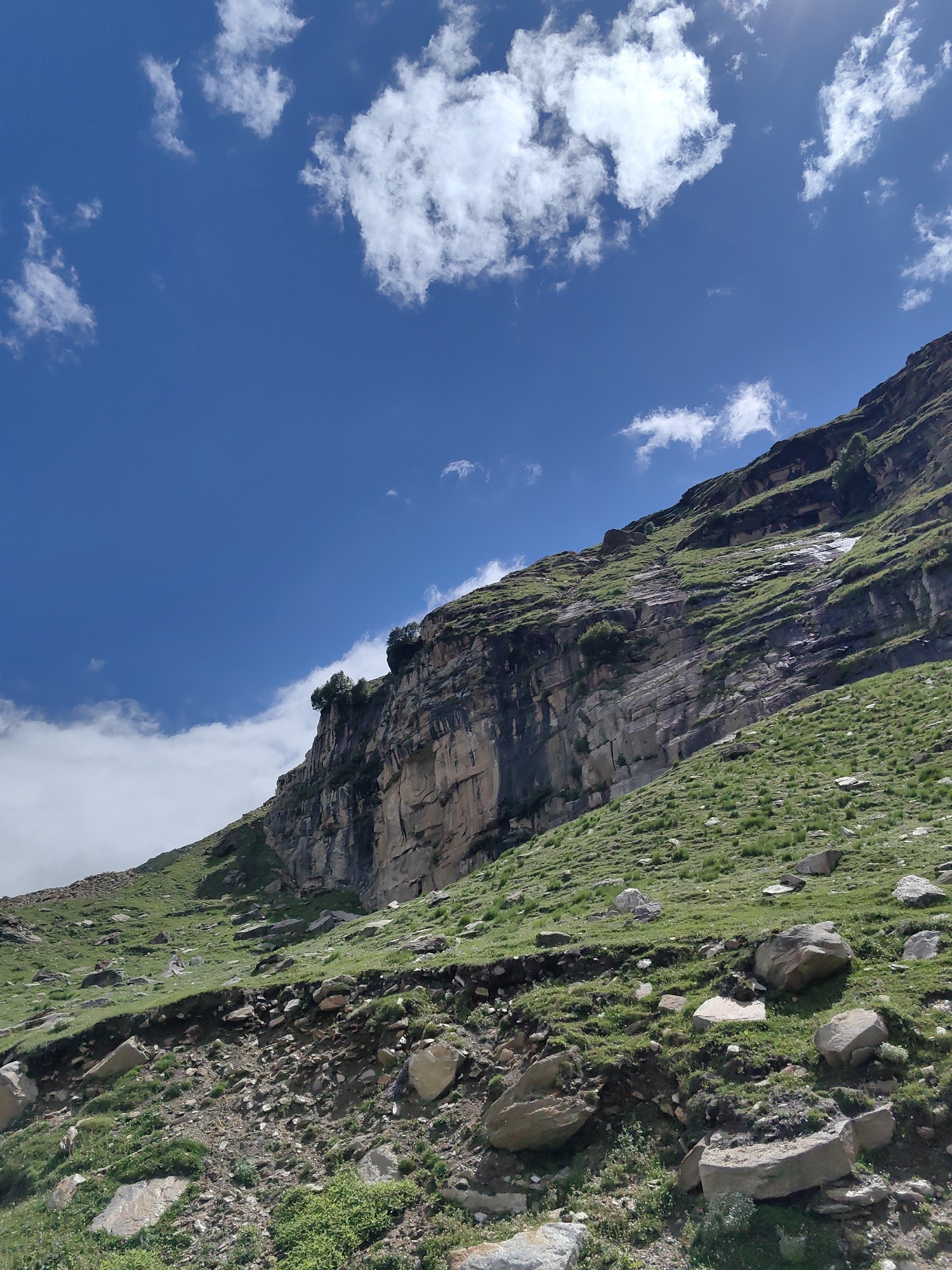 The image depicts a steep, rocky mountainside covered in green grass and scattered boulders under a bright blue sky with scattered white clouds. The sun's rays are visible in the upper right corner, indicating a sunny day. The terrain suggests a high-altitude environment, possibly a remote mountain pass or a slope in the Himalayas. The scene is devoid of any human presence or man-made structures, emphasizing the raw, natural beauty of the landscape. The location is identified as Gramphu, India, a place known for its scenic beauty and mountainous terrain.
