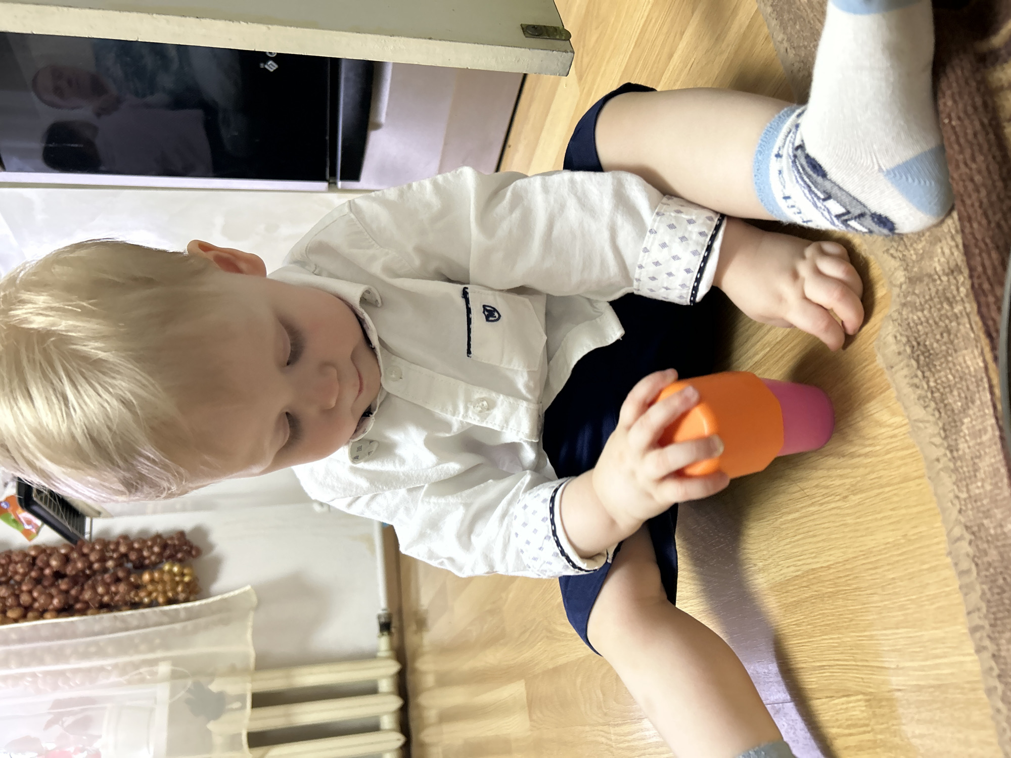 A young, blonde-haired child, identified as a toddler, is seated on a light brown wooden laminate floor in a residential interior in the city of Фоки, Russia. The child wears a white long-sleeved collared shirt with patterned cuffs, dark blue shorts, and white socks with blue accents and a gray vehicle motif on the left foot. The child's gaze is directed downwards, focused on an orange stacking cup held in the right hand, which rests atop a pink stacking cup. A segment of a textured brown rug is visible in the lower right foreground. In the background, a white radiator is present beneath a window draped with a translucent white curtain. To the right of the window, multiple strings of braided brown and golden-brown objects, resembling onions, hang vertically. On the far right, a dark, reflective surface, consistent with an appliance door, displays faint reflections of at least two human figures, one appearing adult. A light-colored doorframe marks the extreme right edge of the scene.