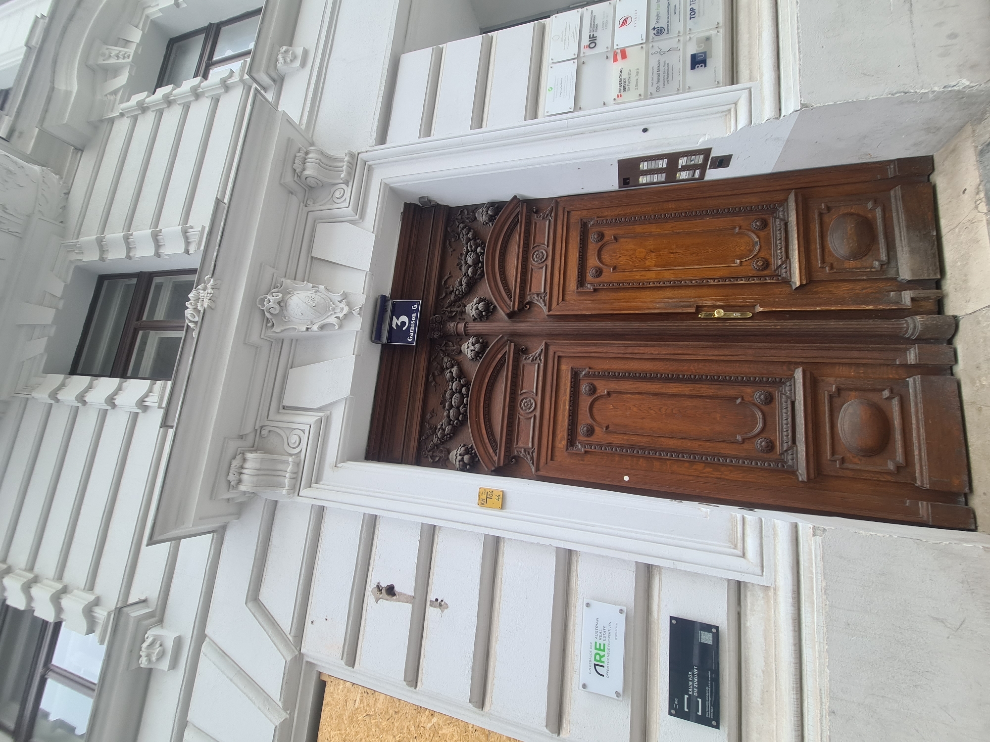 A low-angle, full-shot photograph captures the ornate, dark wood double doors of a building in Vienna, Austria. Above the doors, a rectangular black sign displays the white numerals "3" and "Garnison-G." The building's facade is white and features intricate architectural details, including decorative moldings and reliefs. To the left of the entrance, a sign for "ARE Austrian Real Estate" is affixed to the wall. To the right of the entrance, a series of rectangular white signs are mounted on the facade, with the topmost one displaying "OIF." Further down, other signs are visible, including one with the text "BUG" and another that reads "TOP TE." The doors themselves are made of richly colored wood with carved panels and decorative metalwork, including a golden door handle. The area surrounding the entrance appears to be a street or sidewalk.