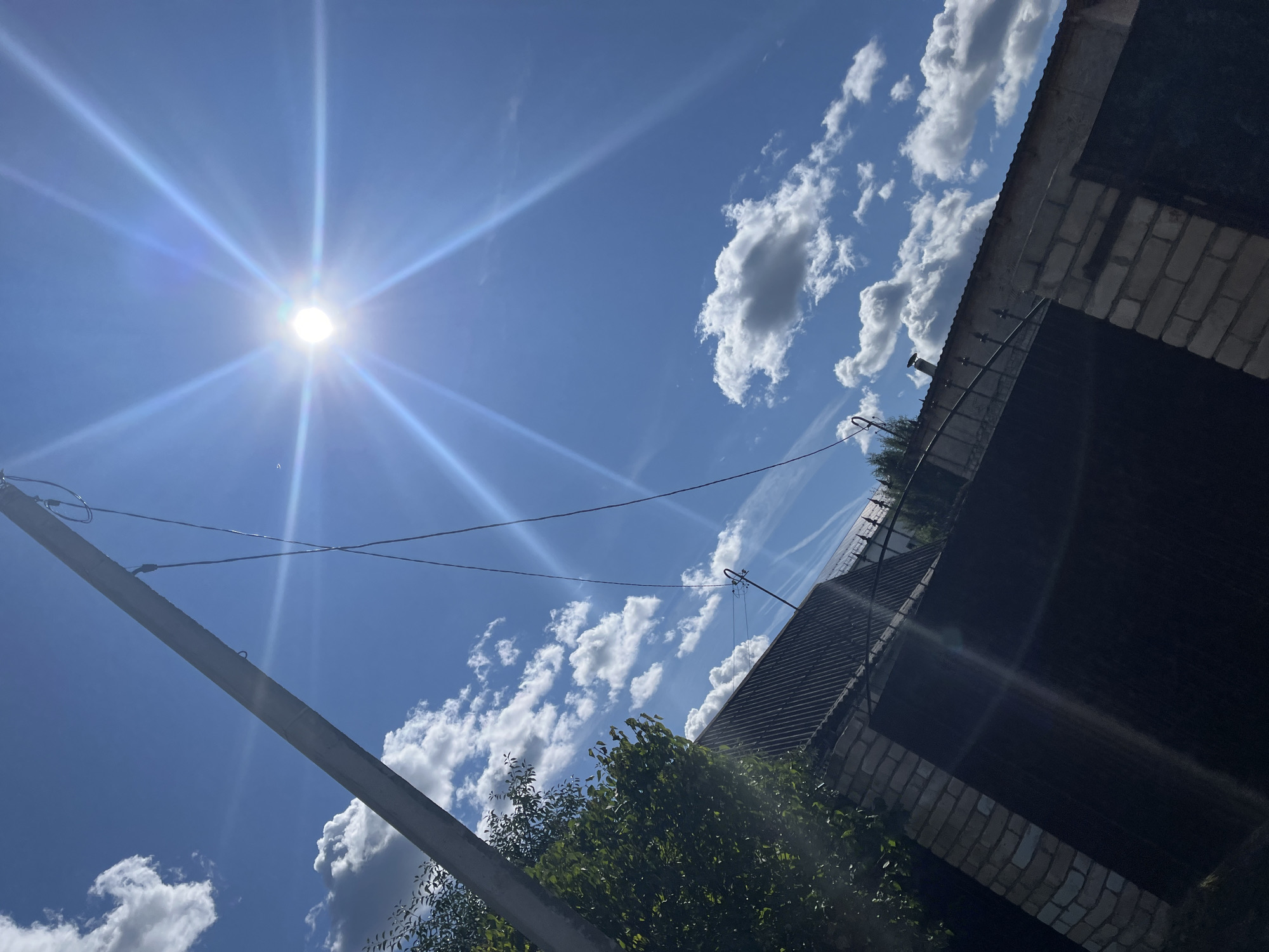 The image depicts a bright, sunny day with a clear blue sky and scattered white clouds. The sun is prominently featured, casting strong rays of light. In the foreground, a portion of a fence made of brick and metal is visible, along with a dark gate. A tree with green foliage is situated to the left of the fence. A utility pole with wires extends diagonally across the sky. The scene appears to be a residential area, possibly a backyard or entryway, with a roofline of a building visible behind the fence. There are no people or animals present in the image. The overall impression is one of a warm, summer day in an urban or suburban environment.