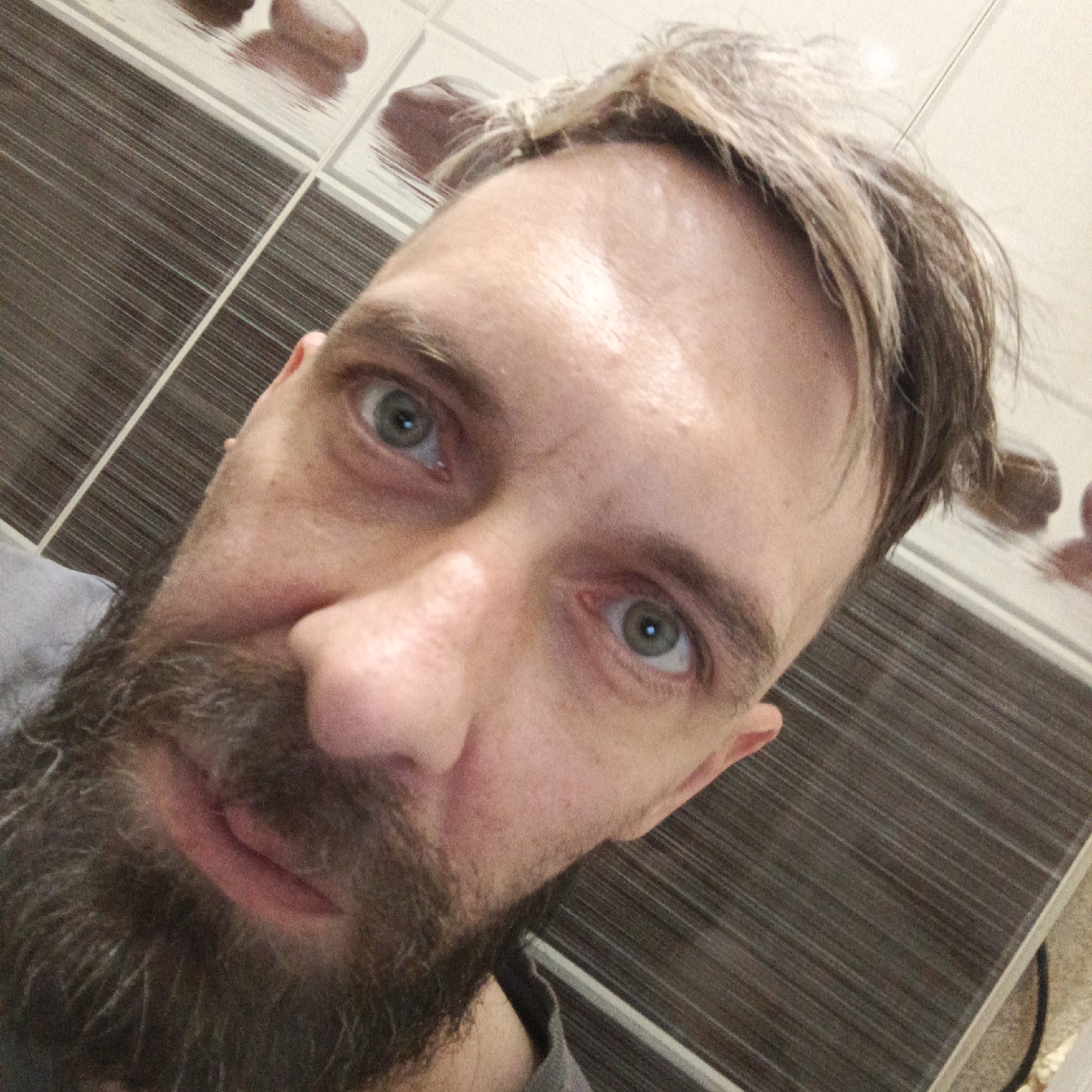 A close-up, upward-angled selfie features a man with a beard and graying hair. The beard is mostly dark with graying strands. His eyes are light-colored, and his skin shows signs of aging with lines and wrinkles. The background consists of a tiled wall with a pattern of alternating white and dark brown stripes, as well as hints of stone-like protrusions. The location of the image is unknown, but has no association with the city of Марковский, Russia.