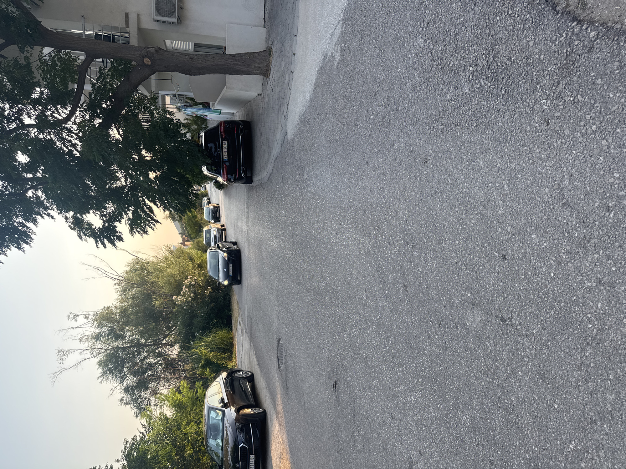 The image captures a street scene in Sutomore, Montenegro, during the late afternoon or early evening, as indicated by the warm, soft light. The perspective is from ground level, looking down a gently sloping asphalt road lined with trees and buildings.
Several vehicles are present on the road. In the foreground, on the left, is a dark-colored sedan, possibly a Skoda, with a license plate that appears to read "3214 BF". Further down the road, a black SUV, likely a Mercedes-Benz, is parked on the right side, its license plate reading "SM1 2018". Other vehicles, including a black hatchback and a silver car, are visible further up the street, suggesting moderate traffic.
The environment is characterized by dense green foliage on the left side of the road and a white building with balconies on the right. A large tree with dark green leaves extends into the frame from the right, partially obscuring the building. The sky is a pale, hazy blue, transitioning to a warmer hue near the horizon, consistent with sunset. The road surface is asphalt, showing signs of wear, and there's a manhole cover visible in the center of the road. A small white wall separates the road from the property on the right. The overall impression is of a quiet residential or semi-commercial street in a coastal town. No people are directly visible in the image.