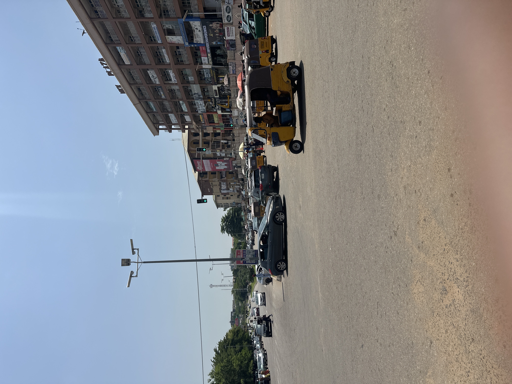 The image shows a busy street scene in Abuja, Nigeria. The orientation is rotated 90 degrees counter-clockwise, making the road appear vertical. On the left side of the road, there is a tall, multi-story building with many windows and balconies. Various advertisements are visible on the building's facade. Further down the street, more buildings and trees line the road.
The road itself is filled with traffic. Several yellow auto-rickshaws, also known as Keke Napep, are prominent, with one in the foreground containing a driver. Cars, including a dark-colored sedan in the mid-ground, are also part of the traffic. The scene depicts a typical day with traffic congestion. A traffic light is visible, showing a green signal.
Several street lights are mounted on poles along the roadside. The sky is clear and blue, indicating a sunny day. The overall impression is that of a lively urban environment. No specific emotions are conveyed, but the scene suggests the hustle and bustle of daily life. Text visible on the building includes "COUTURE, DIFFUSER, WIGS" and "ENERGY". There are also signs for "OPENCART" and other businesses. The time of day appears to be daytime, likely mid-morning or afternoon, given the bright sunlight and shadows.