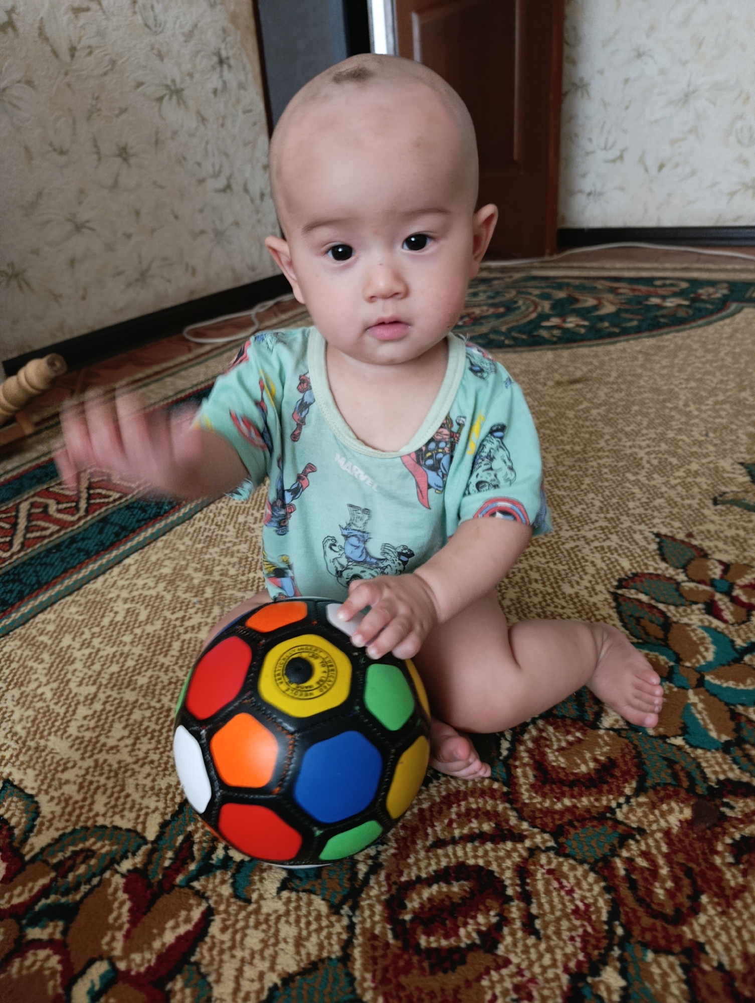 A bald baby sits on a patterned rug in an indoor setting, looking towards the camera with a neutral expression. The baby is wearing a light blue t-shirt with images of superheroes. In front of the baby is a multicolored soccer ball. The background shows a wall with a floral pattern and a closed door. The scene appears to be in a residential environment in Nukus, Uzbekistan.