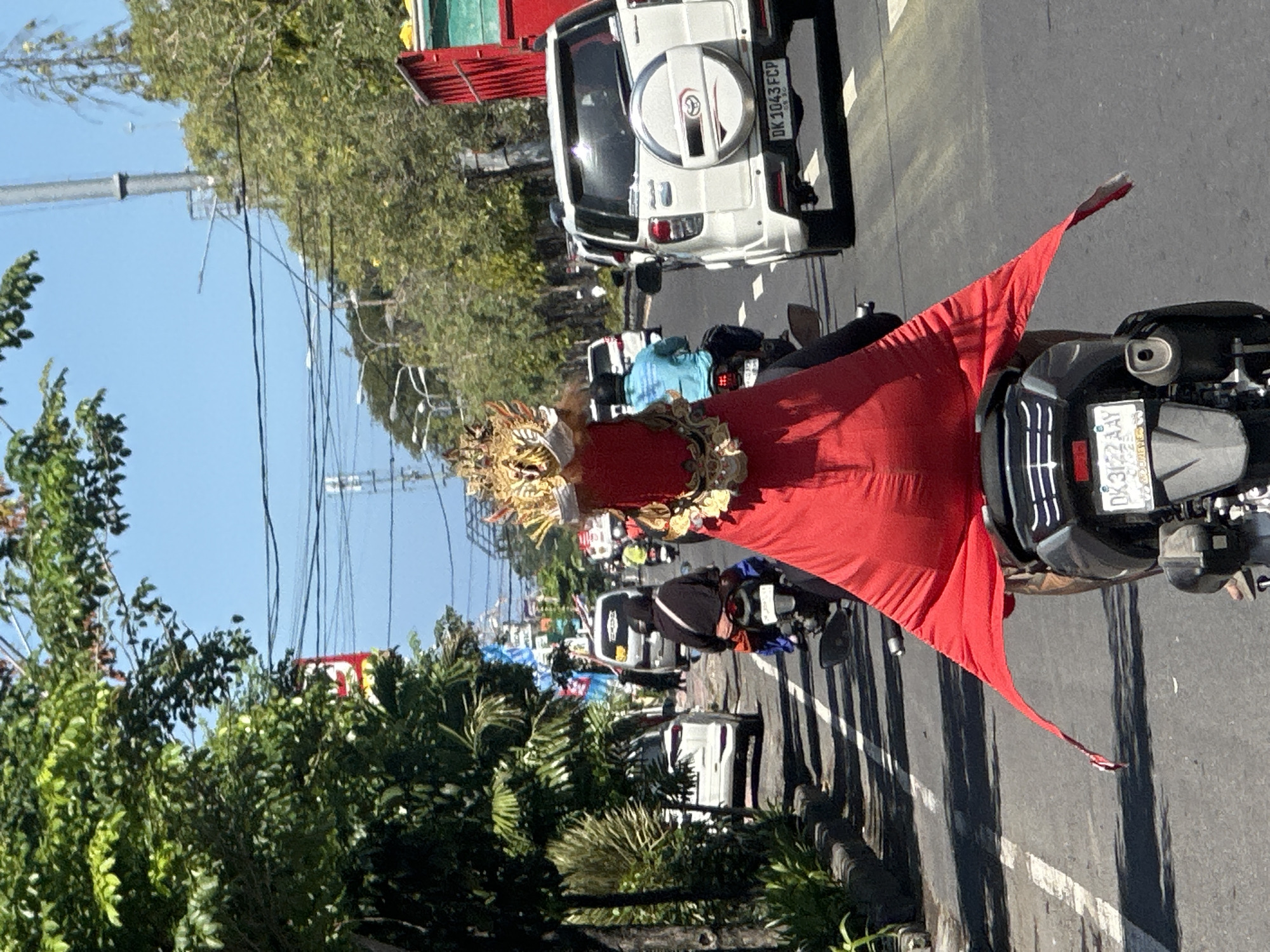 A person wearing a red costume with a gold headdress rides a motorcycle on a street in Denpasar, Indonesia. The person is in the foreground, facing away from the camera, and the costume is draped over the back of the motorcycle. The headdress appears to be elaborate and ornamental, with intricate gold detailing. The motorcycle is dark gray and has a license plate that reads "DK 3122 AAY".
The street is paved and has white lane markings. There are trees and foliage along the side of the road, and several other vehicles are visible in the background, including cars and other motorcycles. The sky is blue and clear. The overall scene suggests a sunny day in an urban environment. The red costume with the gold headdress is the most prominent subject, suggesting a cultural event or performance is taking place.