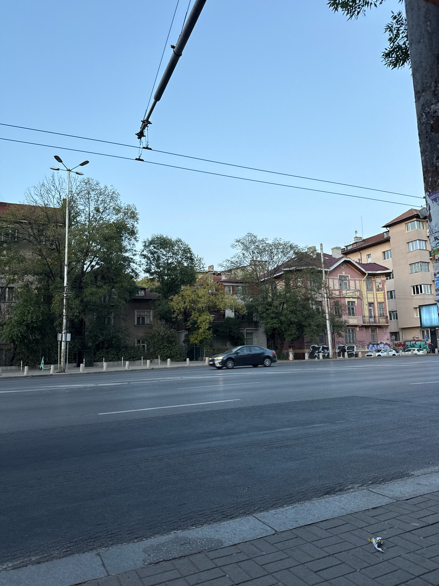 The image shows a street scene in Sofia, Bulgaria. In the foreground, a wide asphalt road runs horizontally across the frame, with white lane markings. The sidewalk on the right is paved with dark grey rectangular bricks. Beyond the road, a sidewalk with a low concrete curb and bollards separates the road from a row of buildings.
Several buildings are visible, varying in architectural style and color. Some are older, darker structures with visible windows and some greenery growing around them. One building in the middle is painted pink and yellow with decorative elements and appears to have graffiti on its lower facade. Another building to the right is taller and lighter beige, with a more modern appearance and multiple windows.
Trees with green foliage are interspersed among the buildings, some of which are quite tall. Overhead, power lines or tram wires are strung across the sky, supported by poles. A street lamp with multiple heads is also visible on the left side of the image.
A dark-colored sedan is driving on the road, moving from left to right. There are a few other cars parked or stopped in the distance near the buildings. A small pile of debris, possibly trash, is on the sidewalk in the lower right corner. The sky is clear and blue, suggesting it is daytime. The overall impression is of a typical urban street with a mix of older and newer architecture.